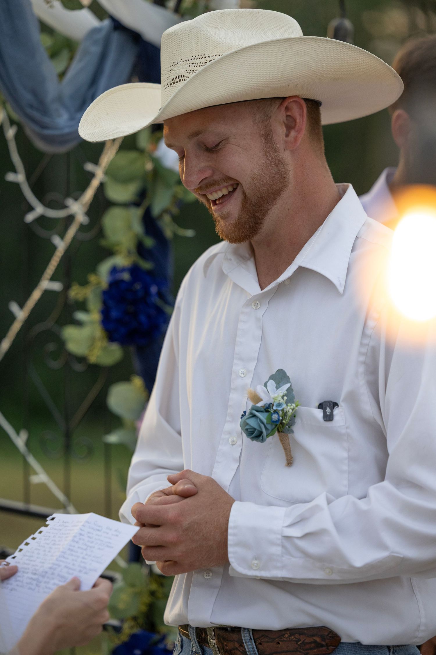 Smiling groom in white shirt and cowboy hat with blue boutonniere at outdoor wedding ceremony.