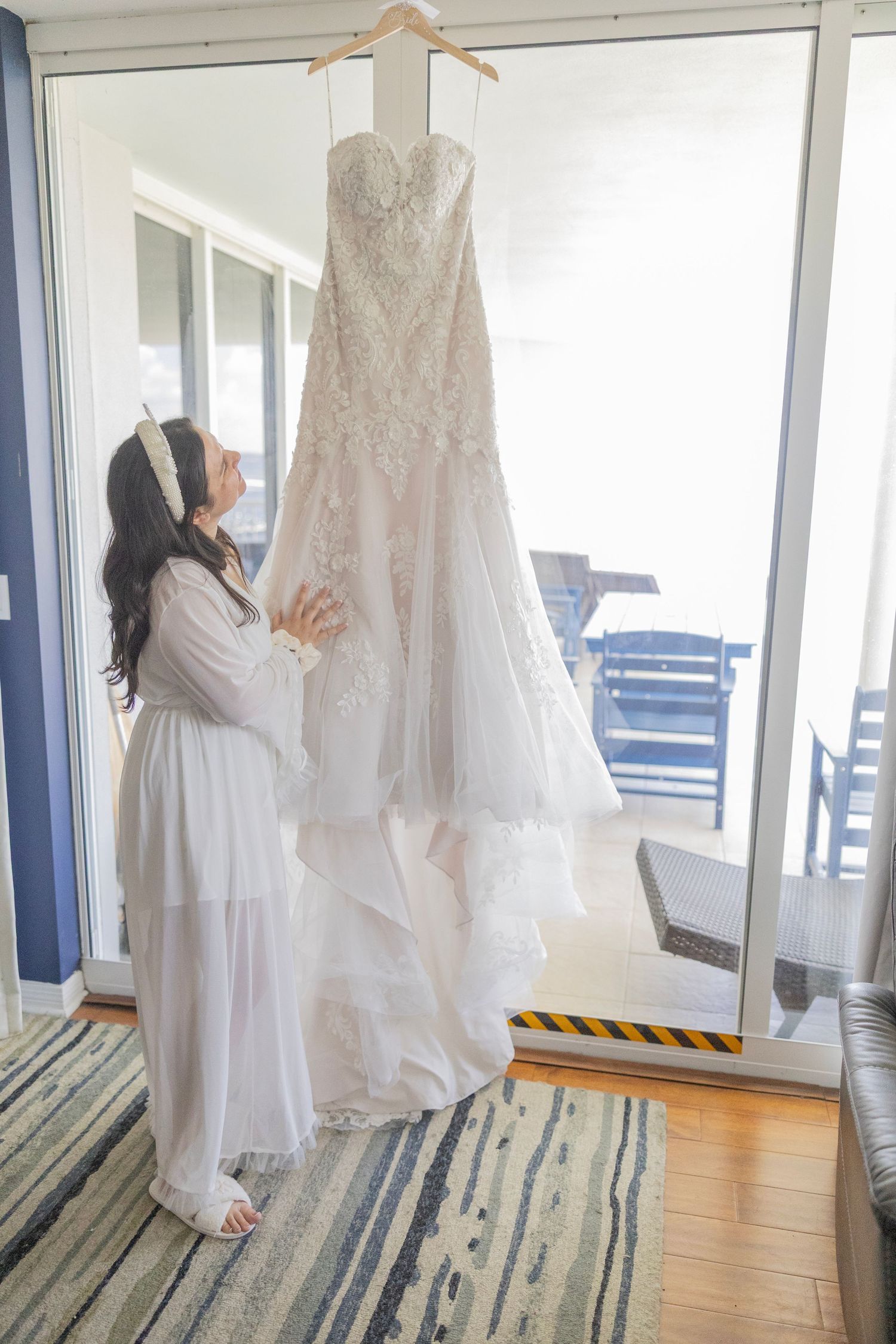 Bride in white robe admiring her lace wedding gown hanging by a sunlit balcony door before her ceremony.
