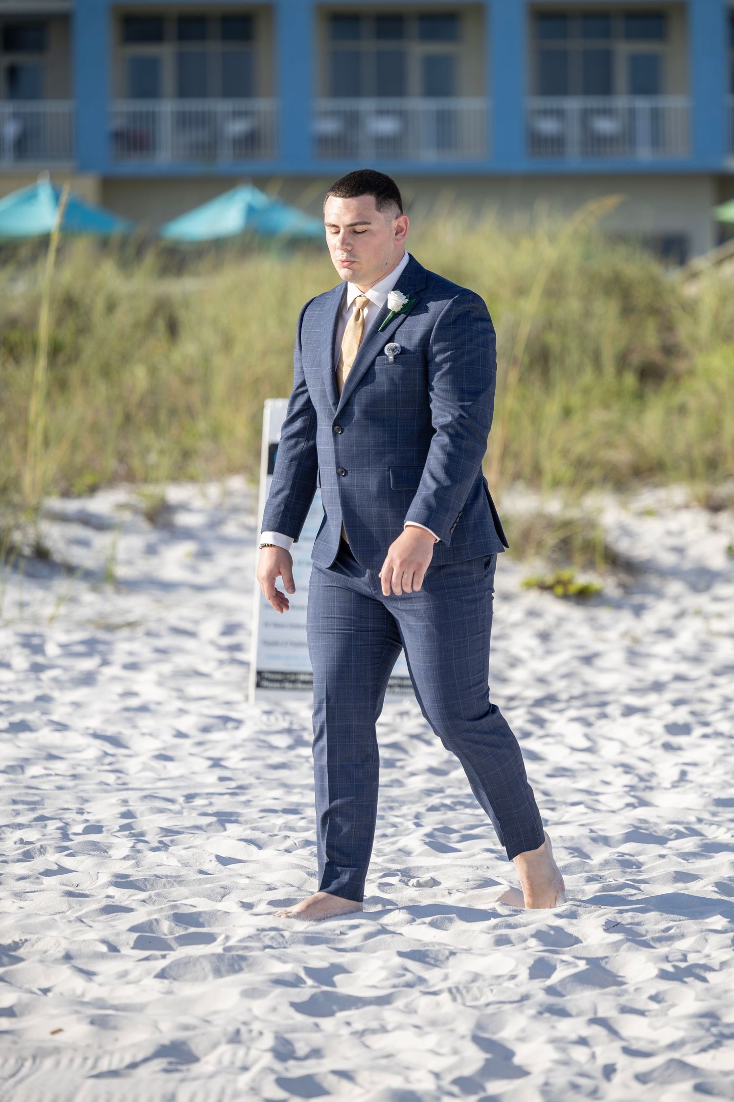 A well-dressed groom in a navy blue suit walks barefoot on white sand beach with a hotel and dunes in the background.