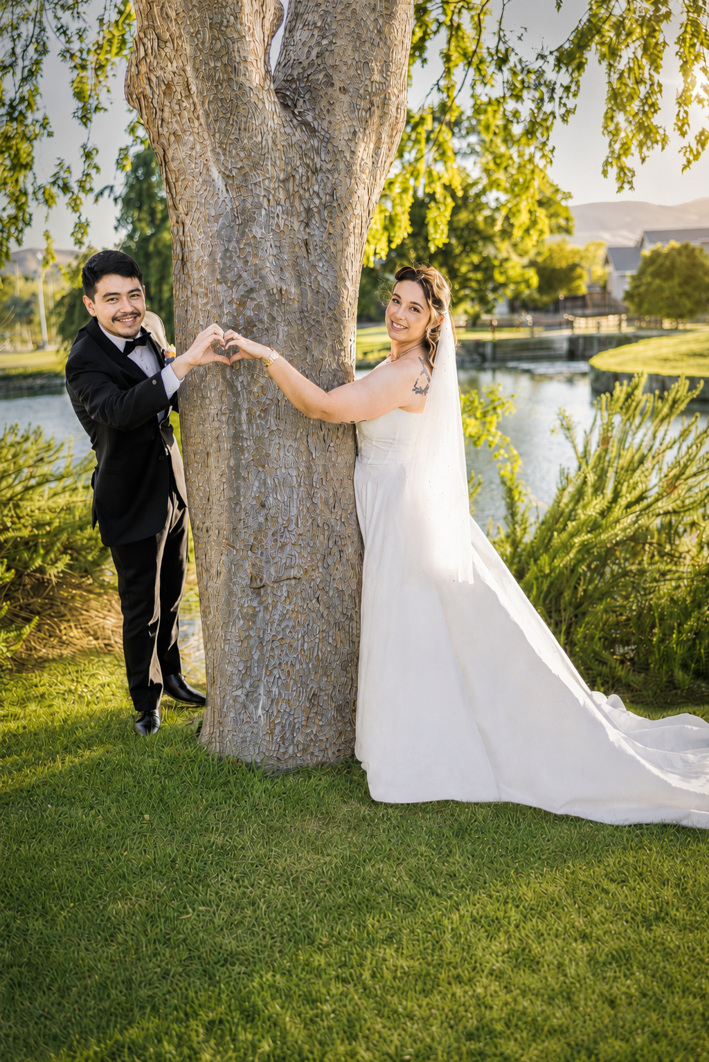 Bride and groom making a heart shape with hands around a tree by a pond on their wedding day.