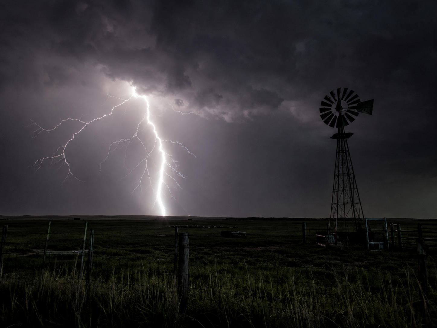 Lightning bolt strikes near a windmill on a dark stormy night in a rural field.