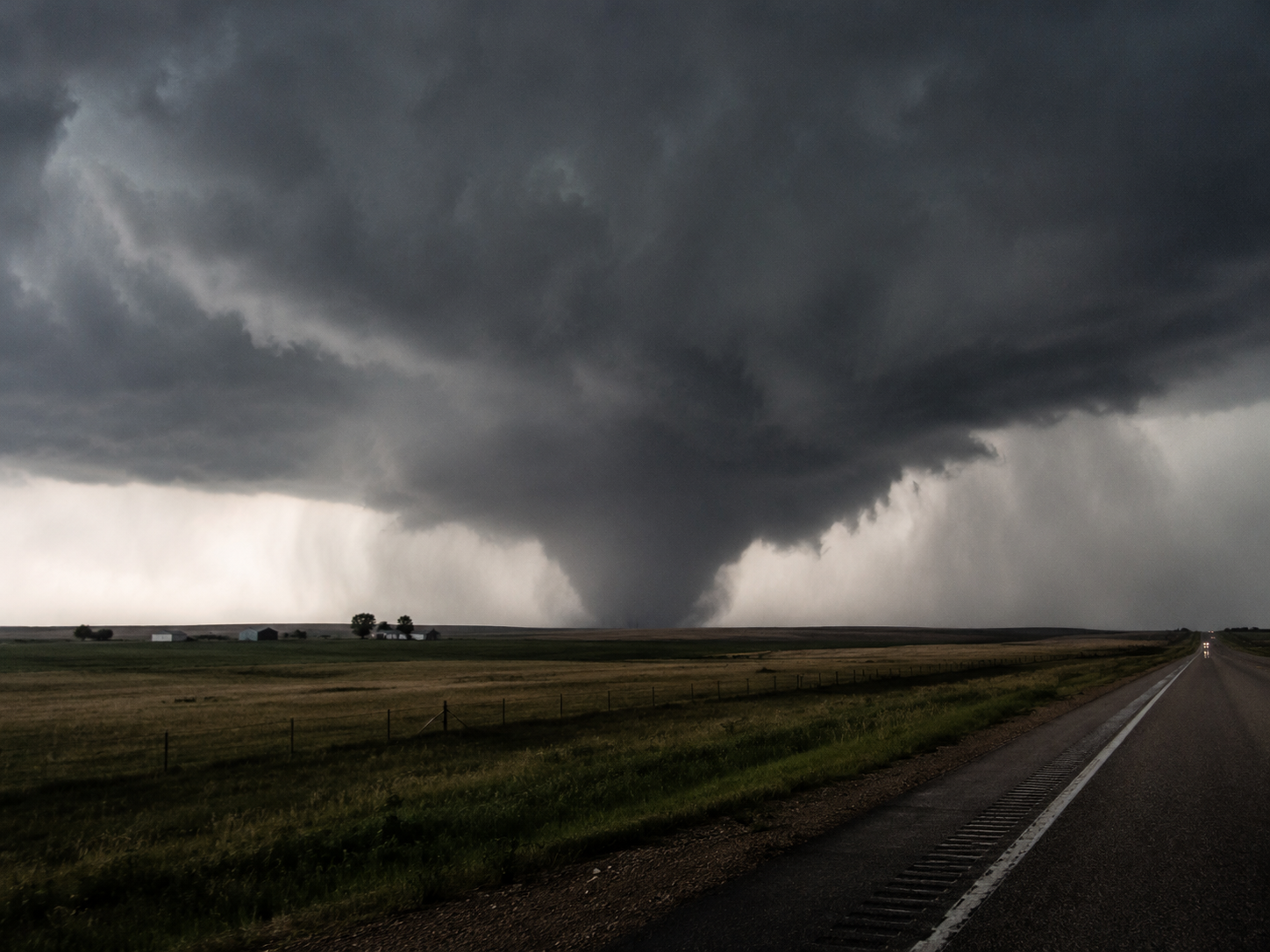 A massive tornado funnel cloud descends over flat farmland fields beside a rural highway under dark stormy skies.