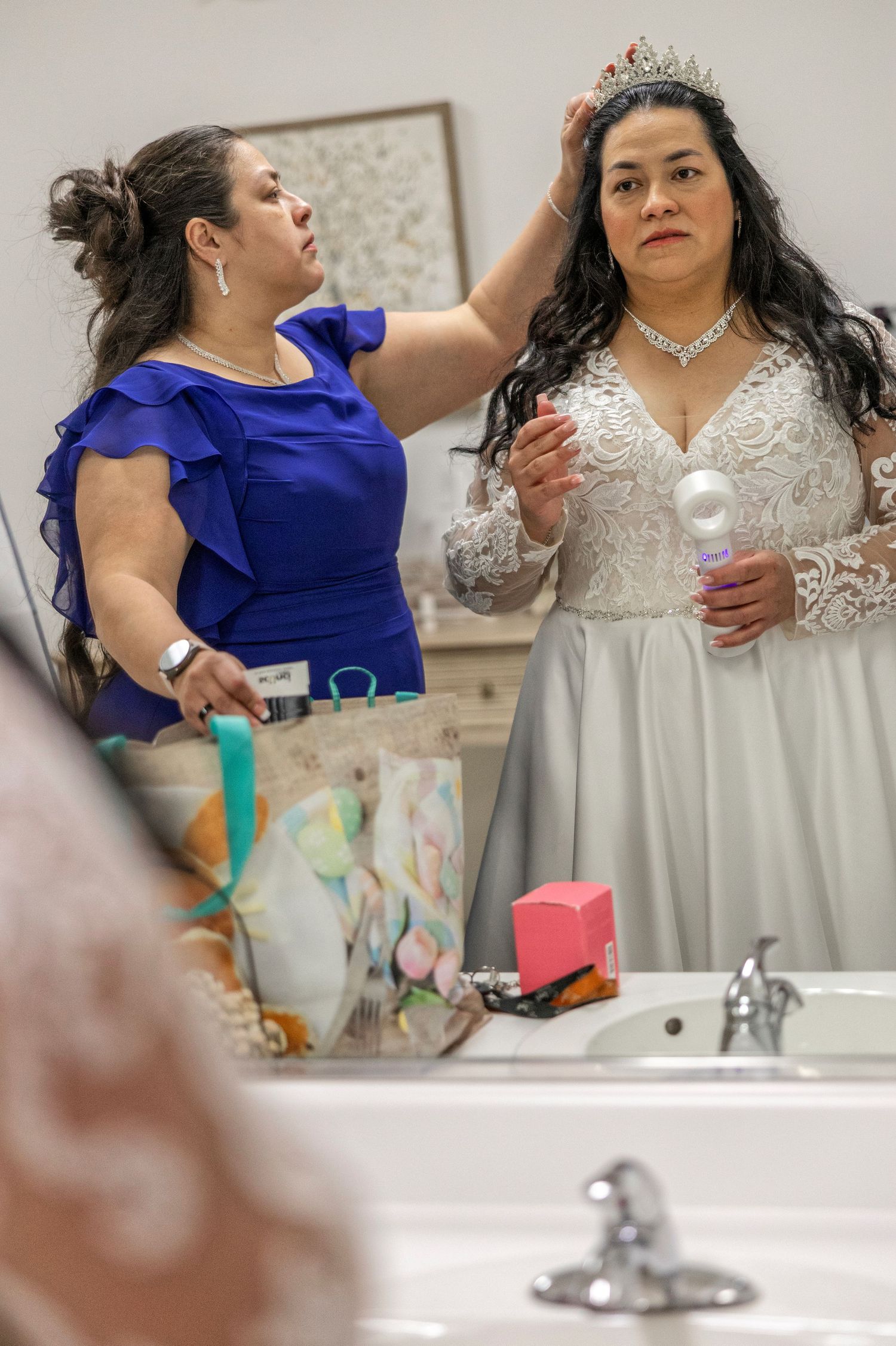 Woman in blue dress adjusts tiara on bride in white lace gown before wedding ceremony in dressing room.