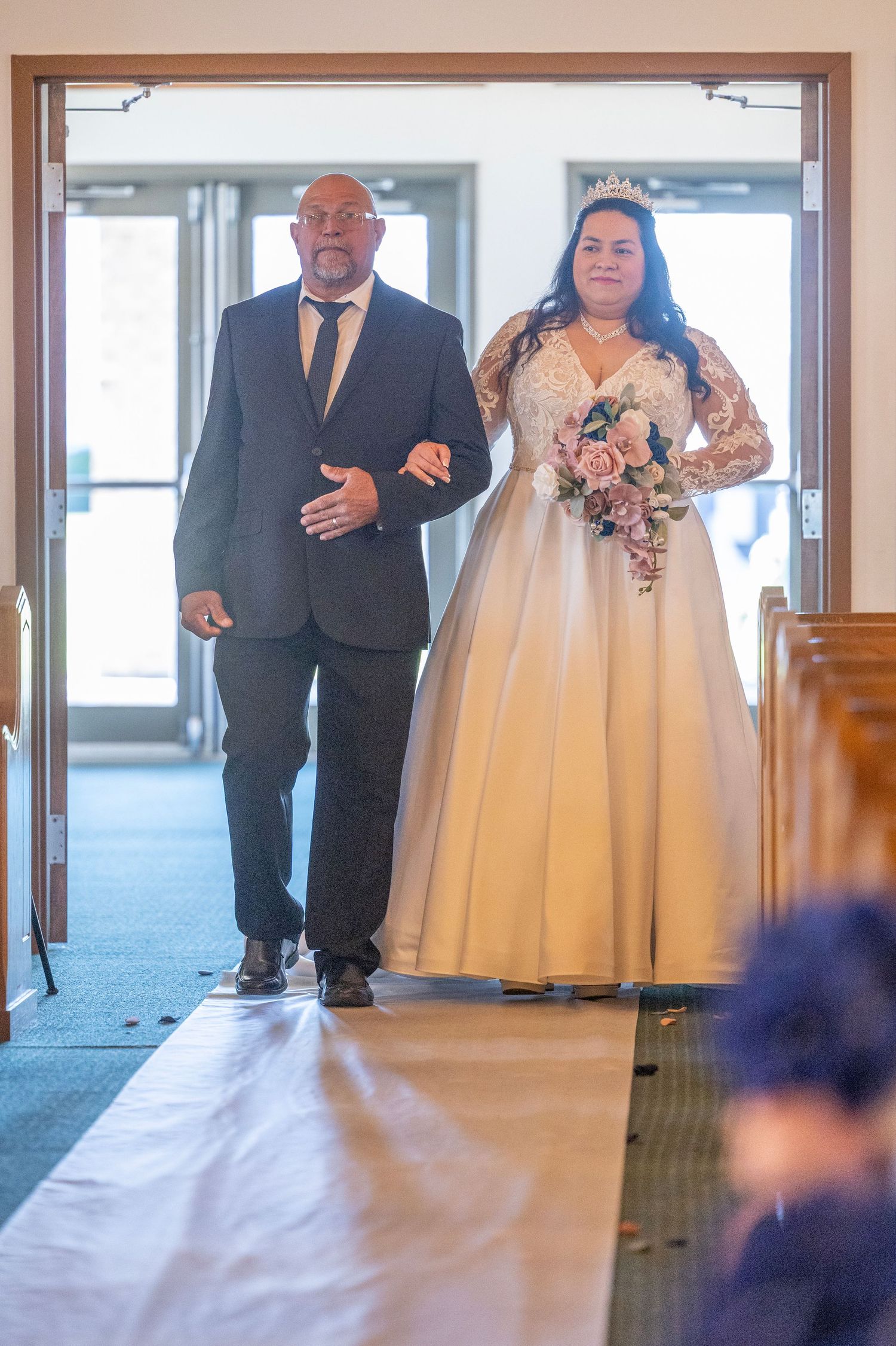 Bride in white lace gown walks down church aisle with escort, holding pink bouquet during wedding ceremony.
