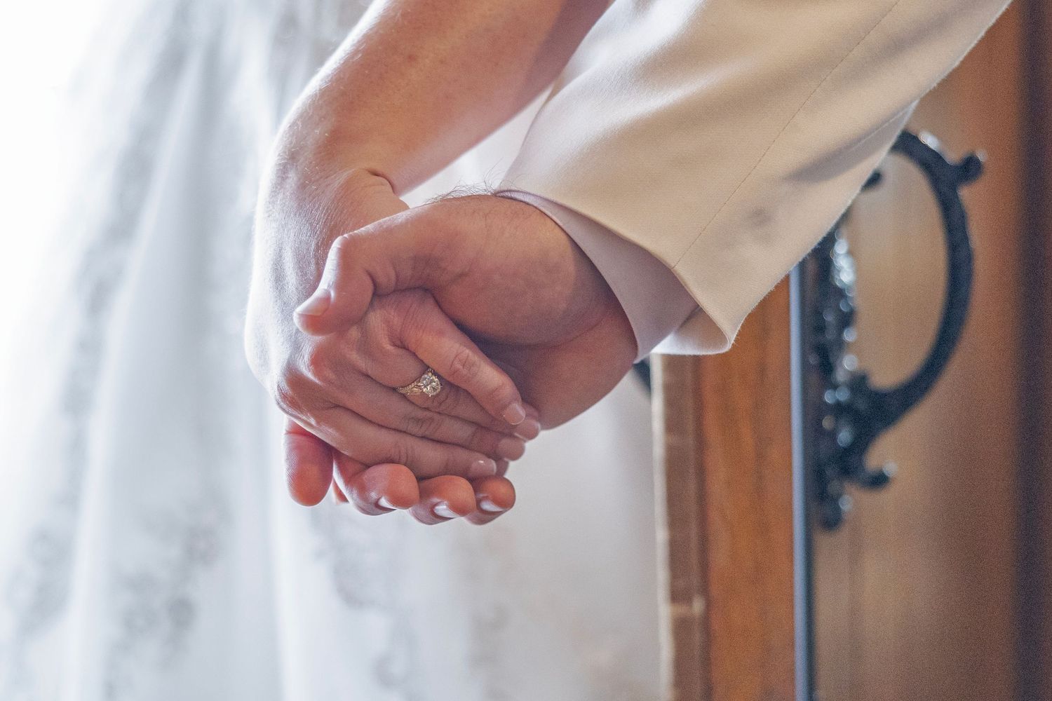 Bride and groom holding hands at wedding ceremony, close-up of rings and white dress.