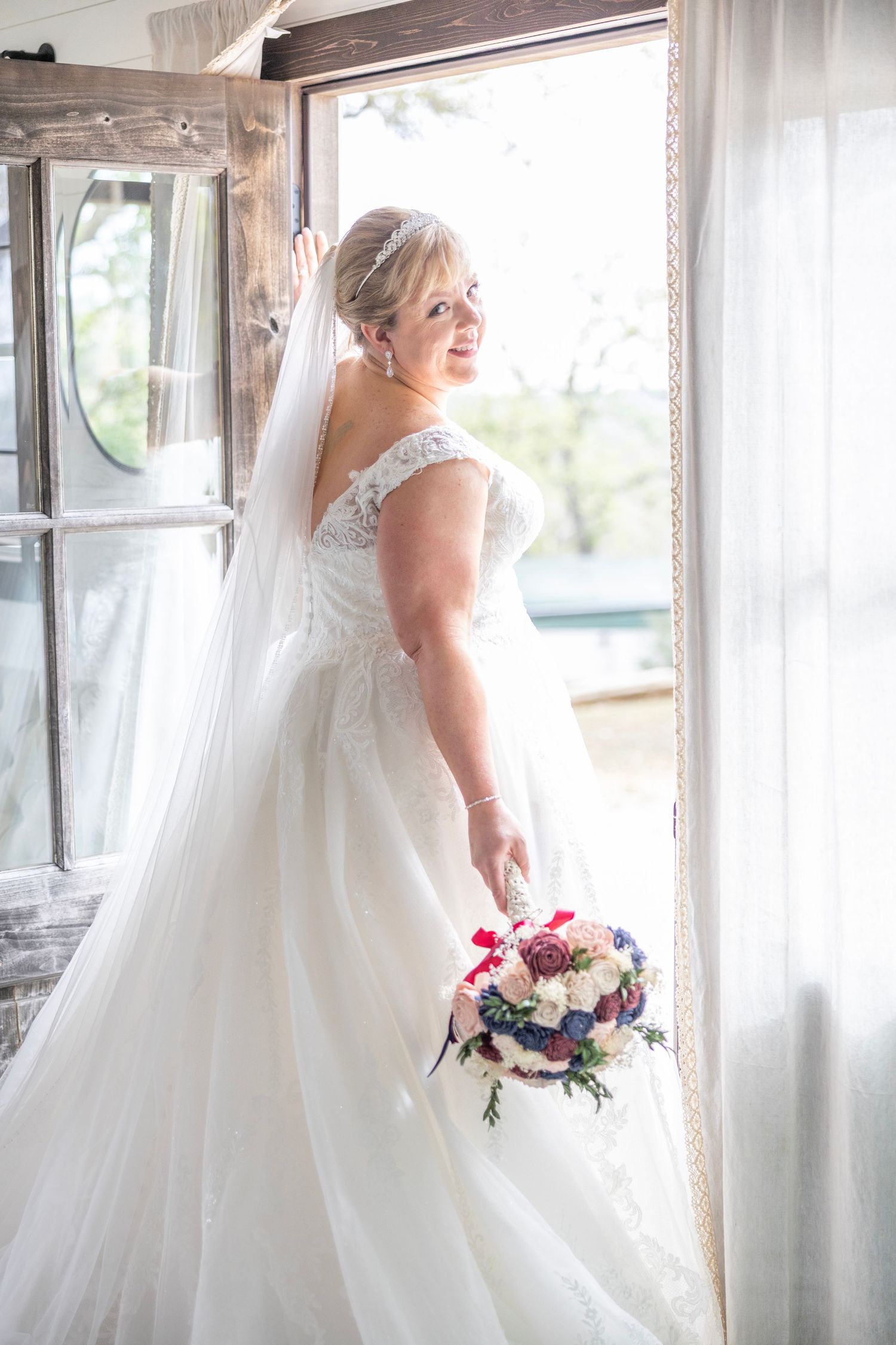 Bride in off-shoulder lace wedding gown with veil holds colorful bouquet, posing by bright window.