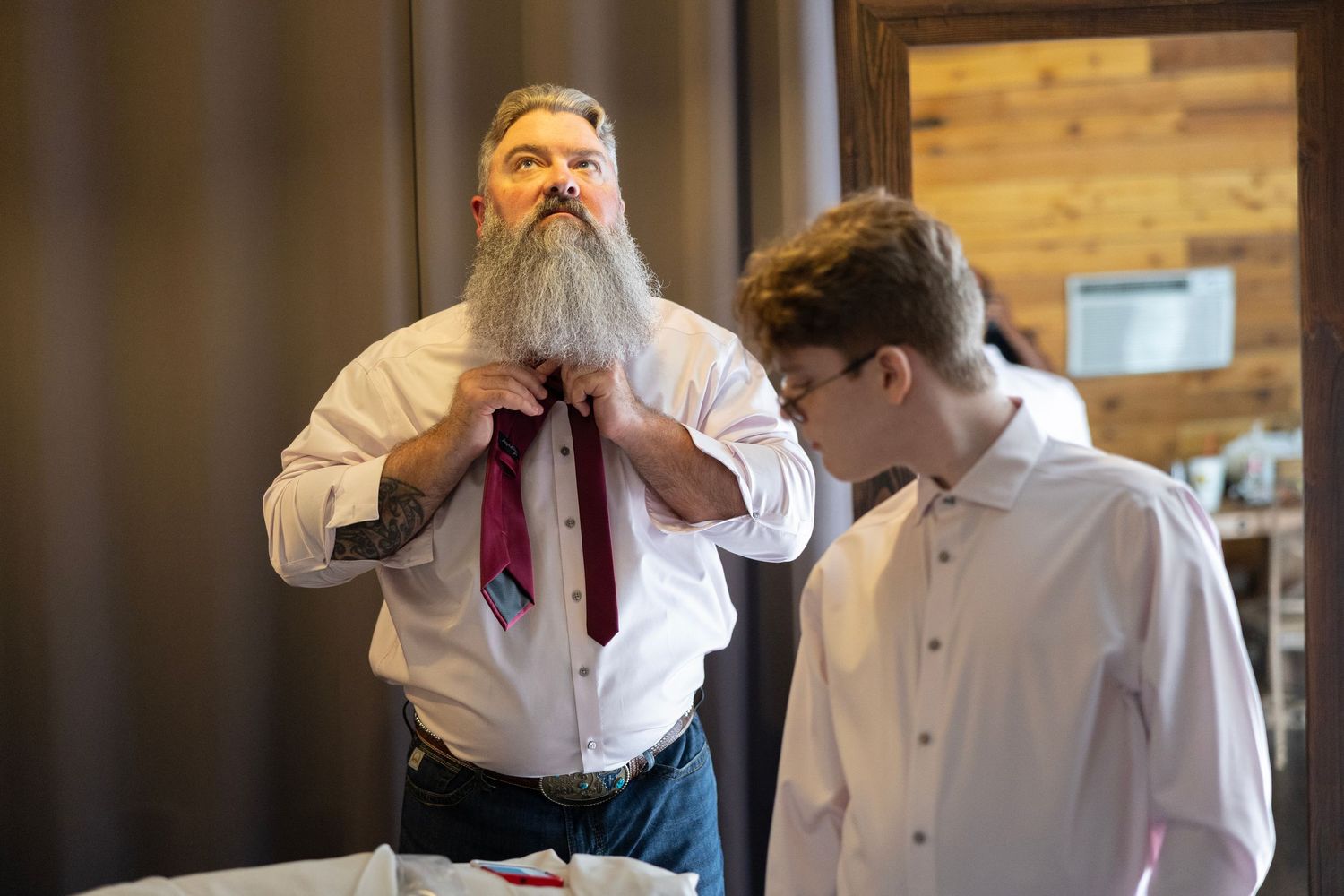 Bearded man in white shirt tying a red tie while a young man in formal wear assists him indoors.