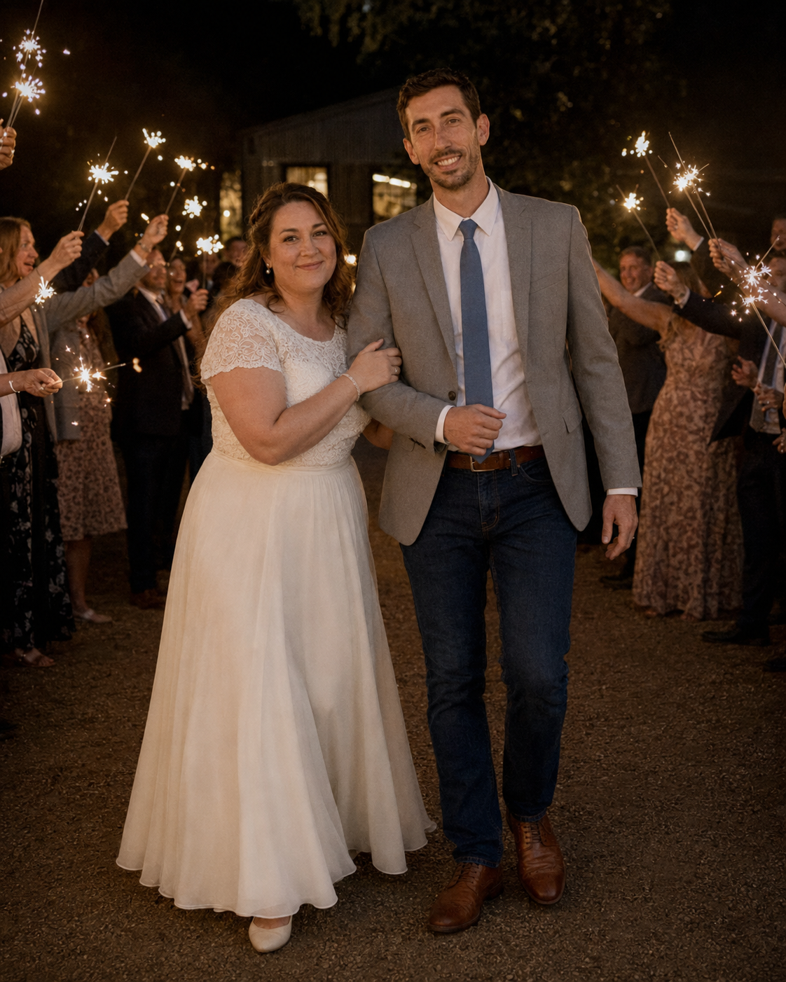Newlyweds walk through sparkler send-off tunnel surrounded by cheering guests during outdoor evening wedding reception.