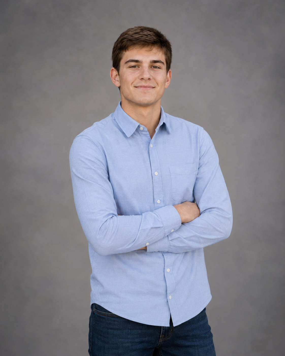 Young man in light blue button-up shirt standing with arms crossed, smiling confidently against gray backdrop.