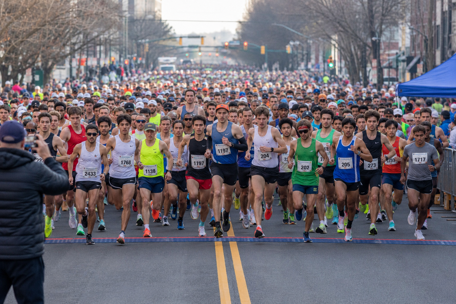 Large crowd of runners starting a road race on a city street, with spectators lining the course on a cool day.