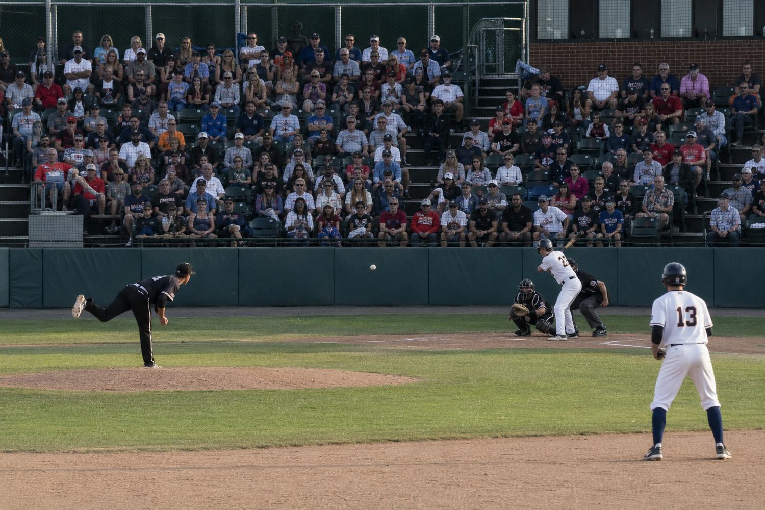 Pitcher delivers pitch to batter while runner leads off first base in night baseball game.