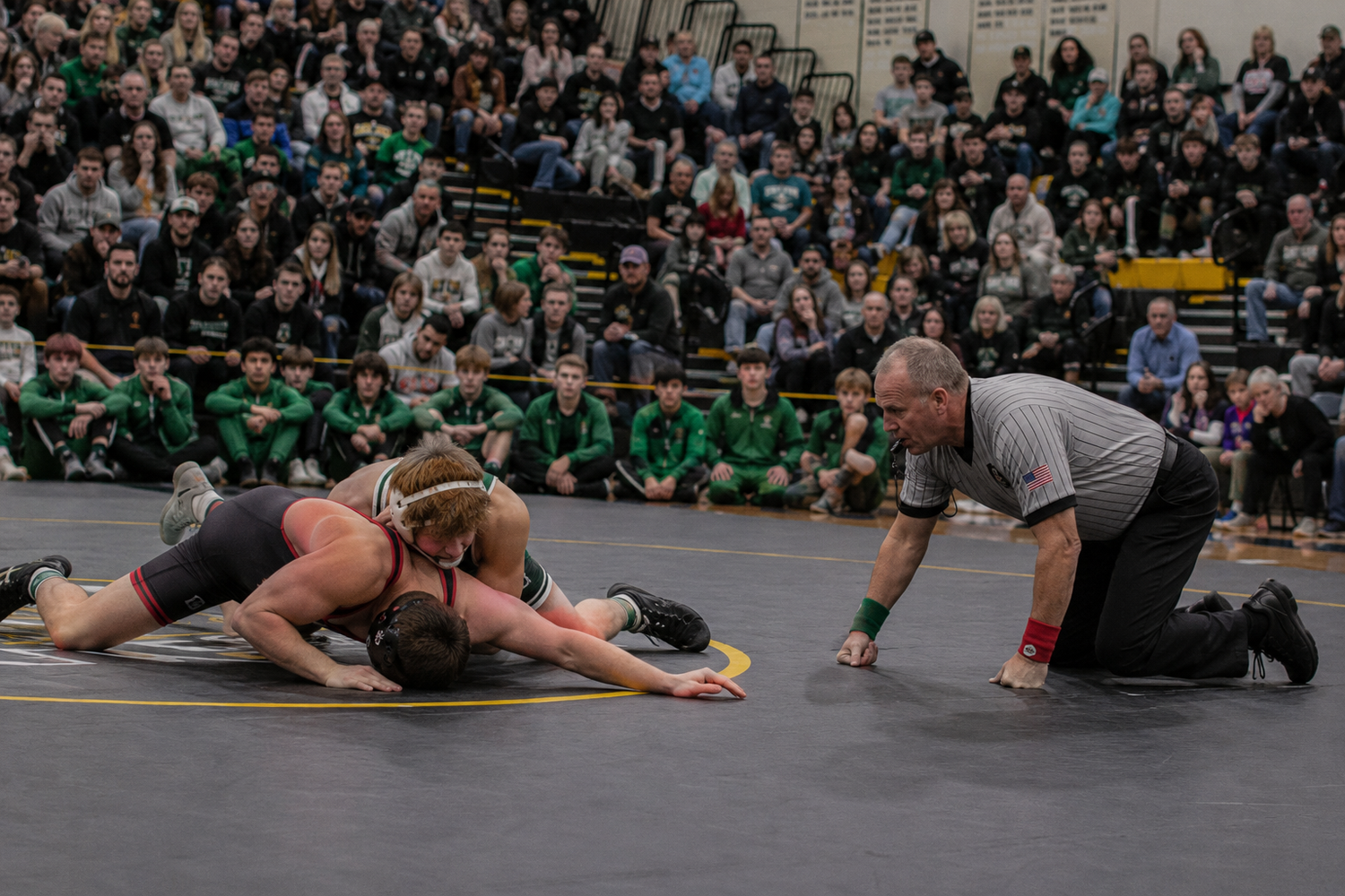 Referee watches closely as two wrestlers compete on the mat during a packed high school wrestling match.