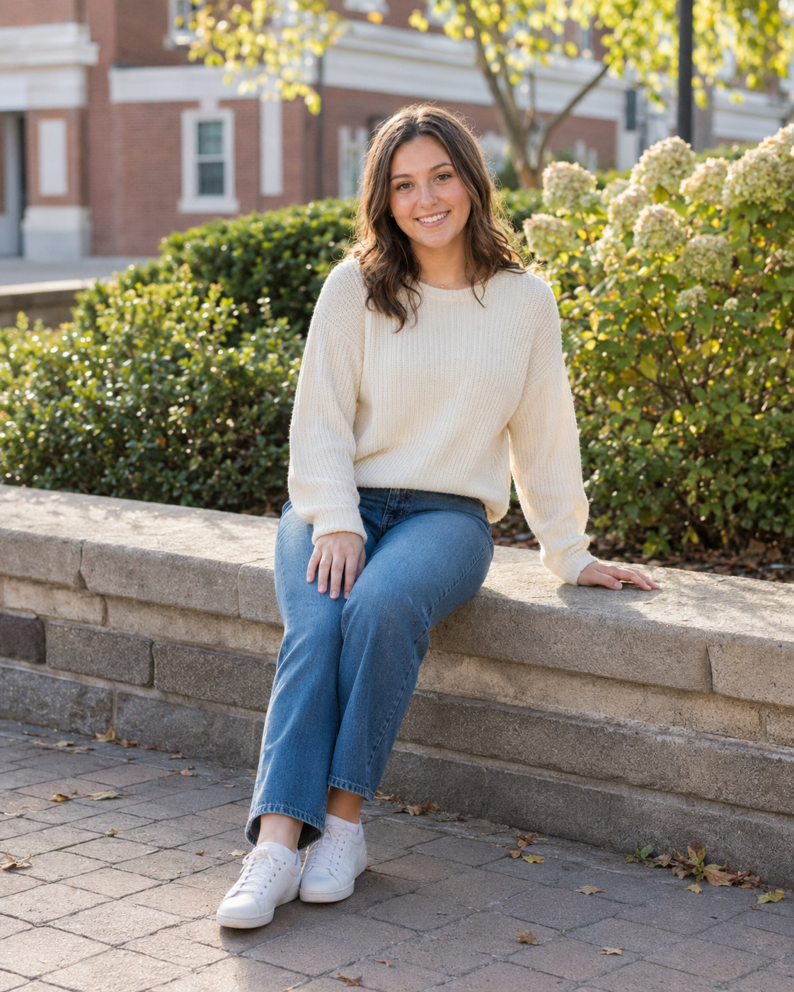Young woman in cream sweater and jeans sitting on stone wall outdoors, smiling in a campus setting.