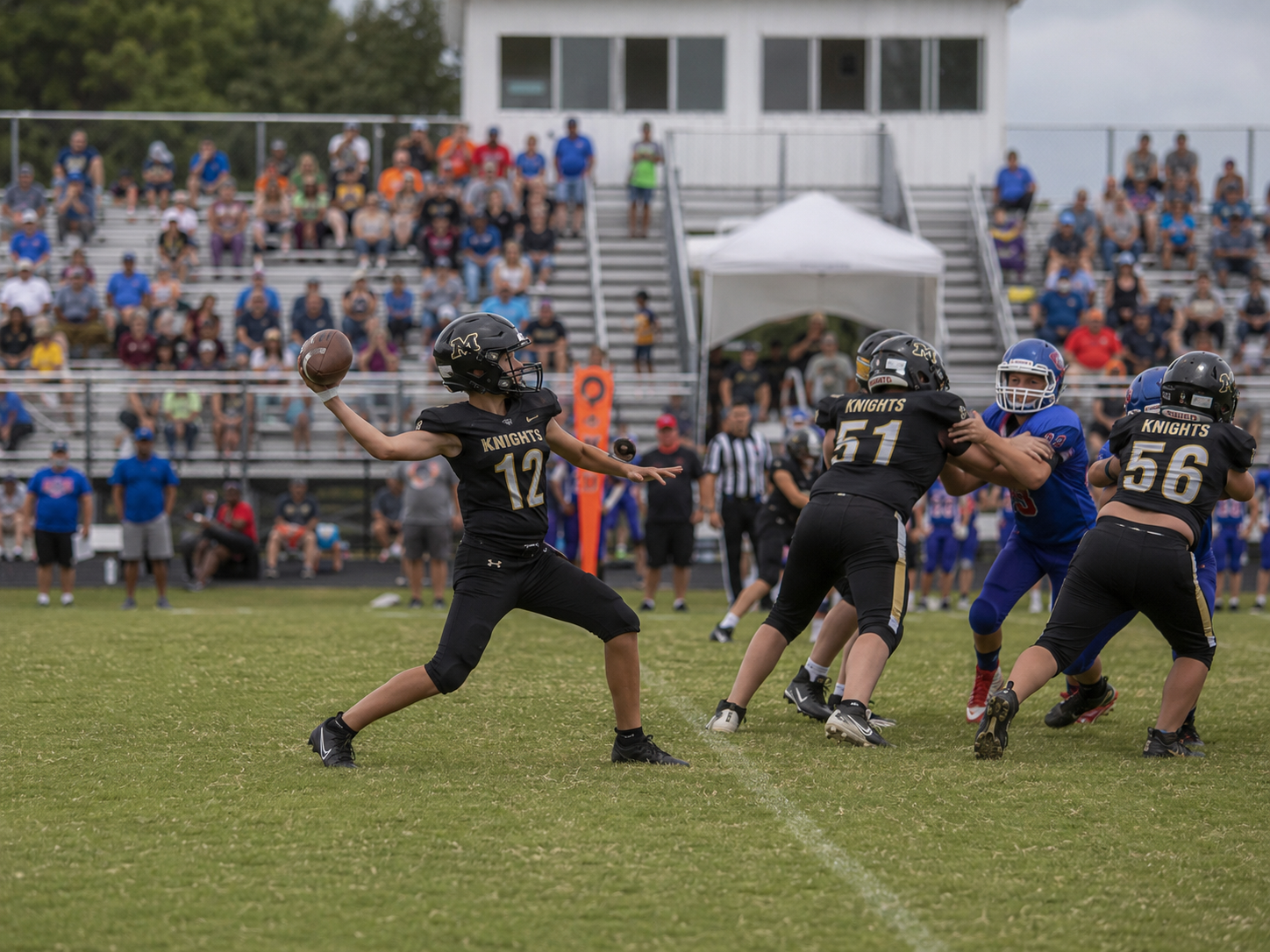 Youth football quarterback wearing number 12 prepares to throw a pass during a game with fans watching in the stands.