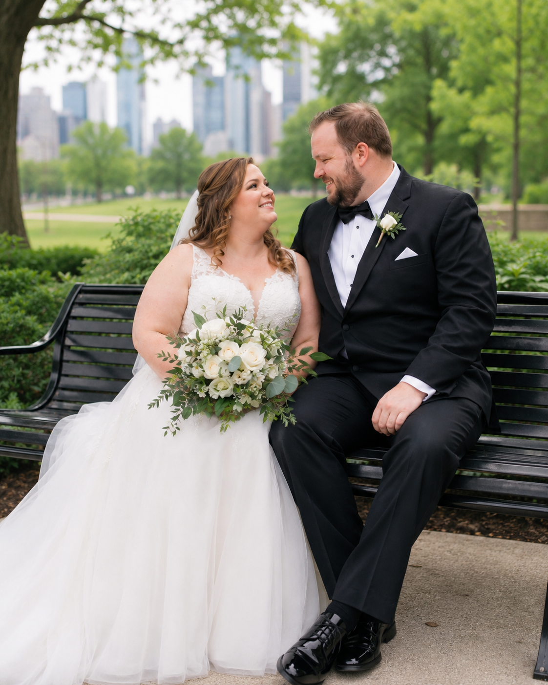 Bride and groom sit on a park bench, smiling at each other, with a city skyline backdrop and white floral bouquet.