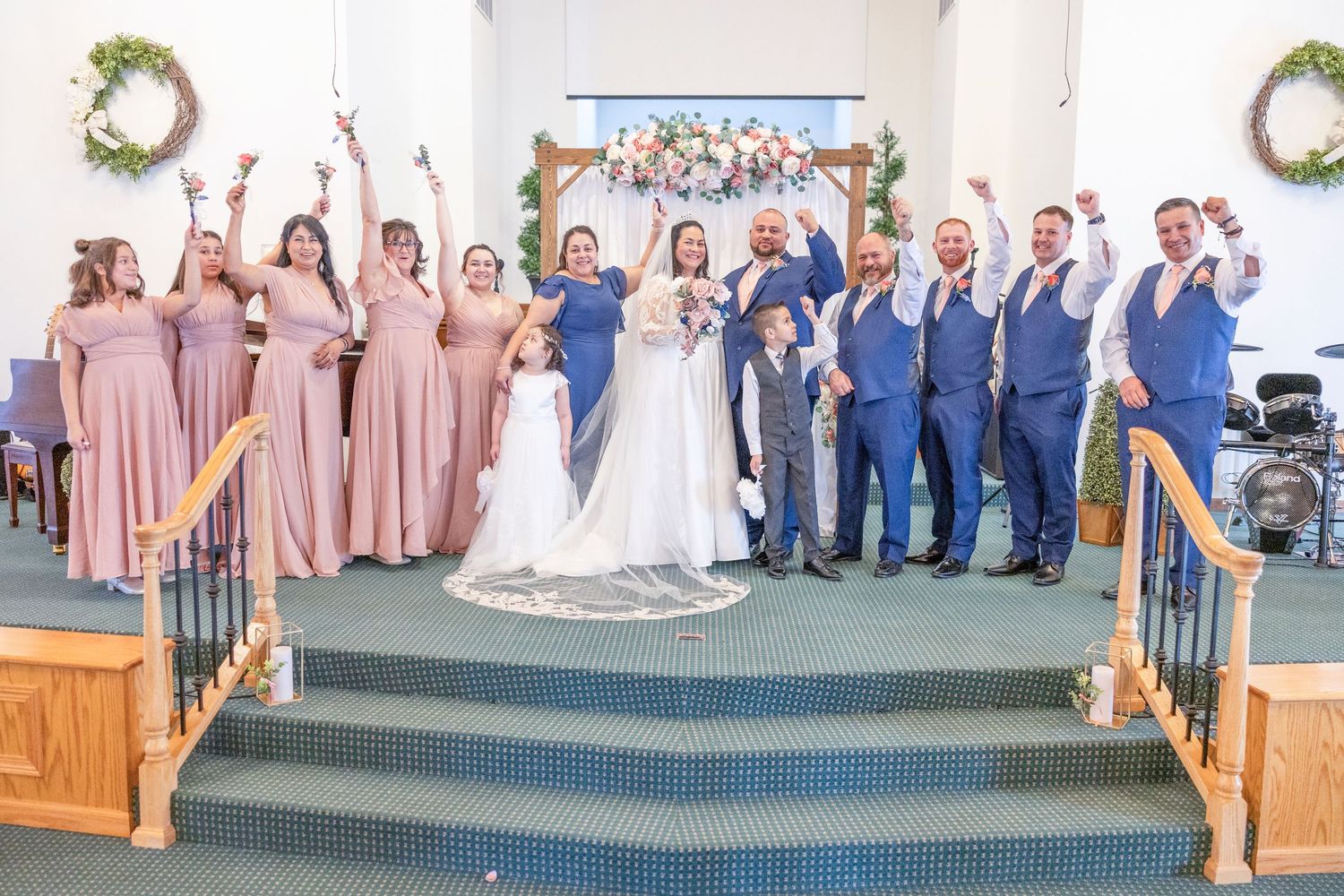 Wedding party celebrating at altar in church, bridesmaids in dusty rose gowns and groomsmen in blue suits cheering with raised arms.