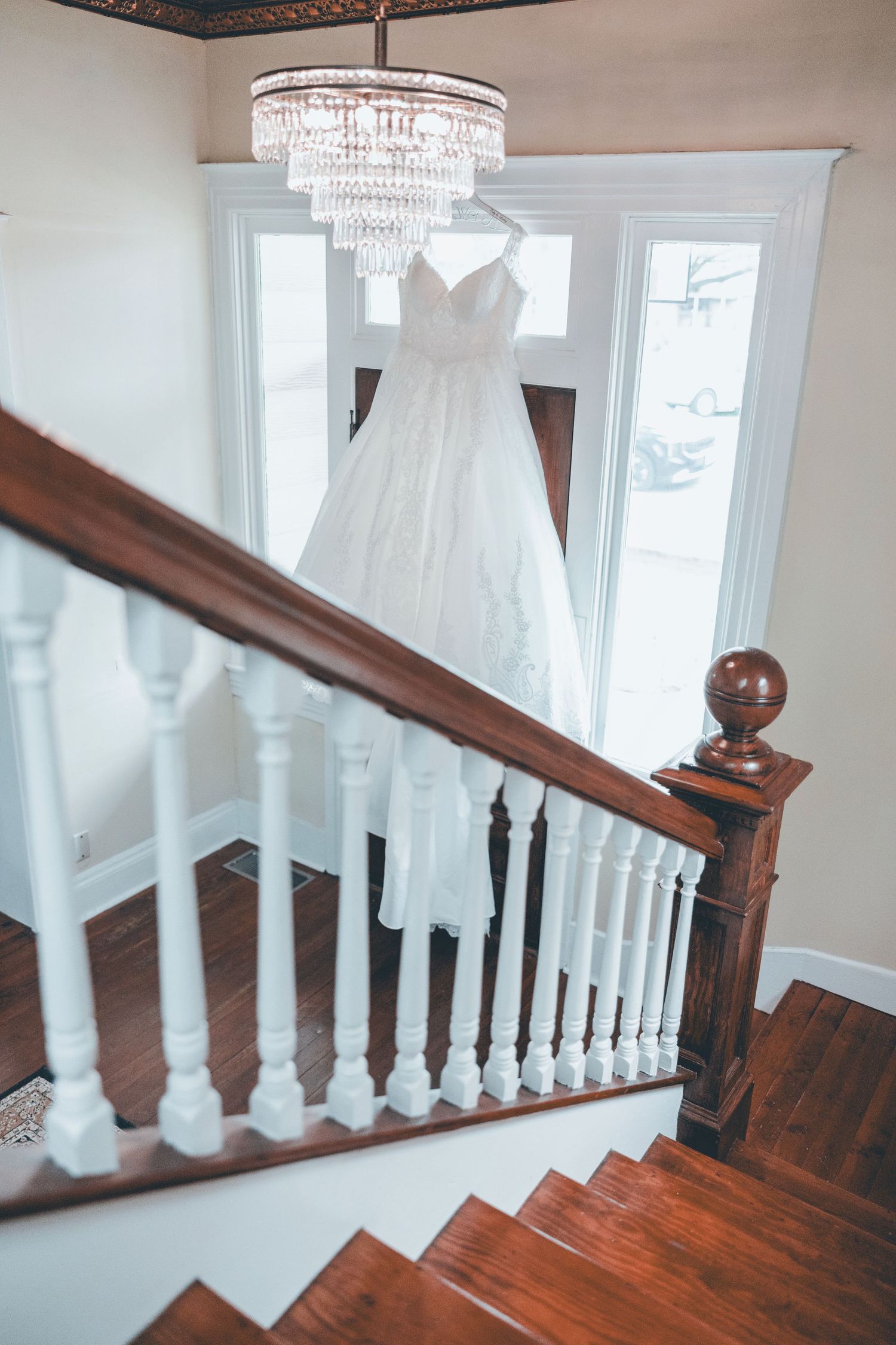 White wedding gown hanging from crystal chandelier above wooden staircase with white balusters in elegant home interior.