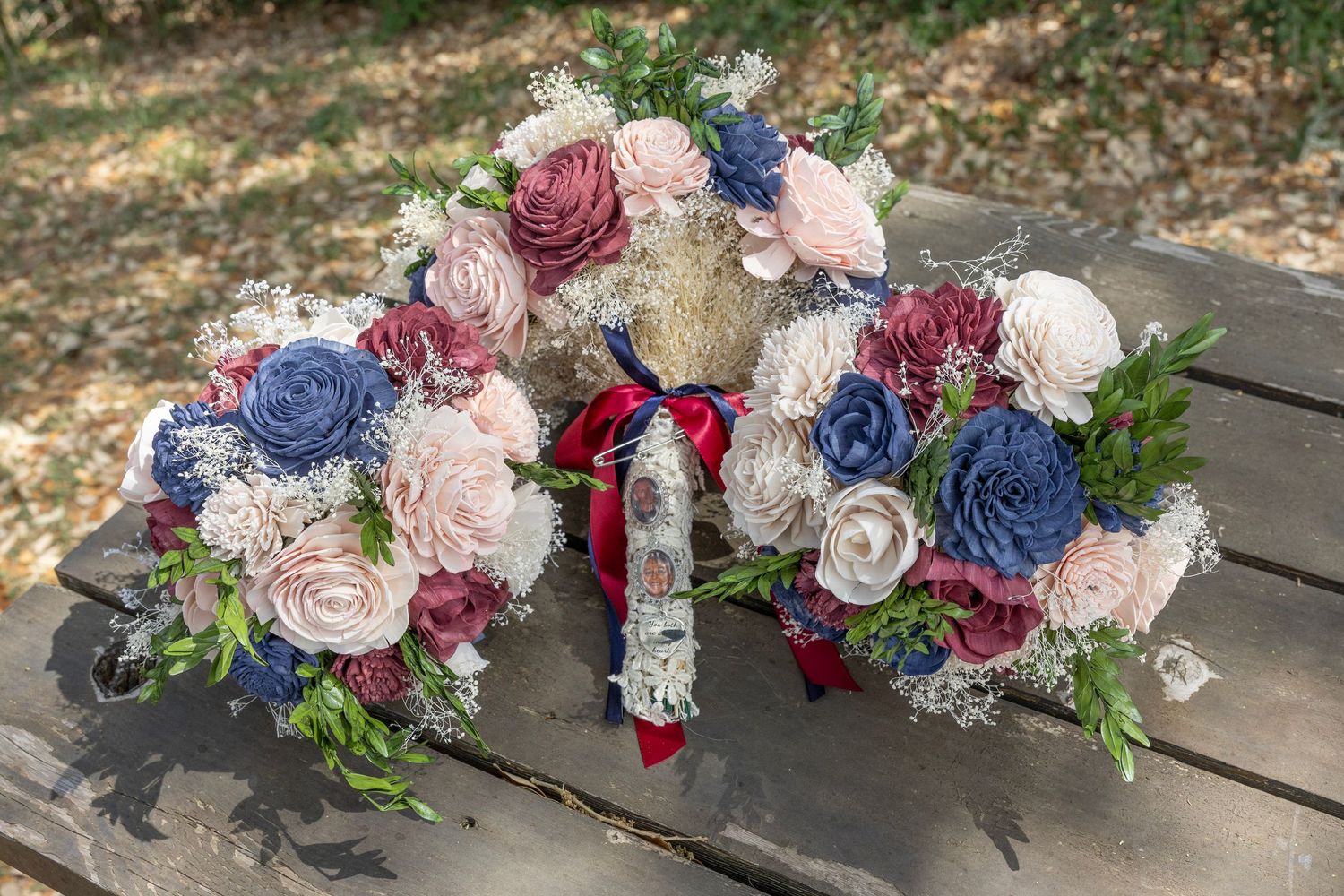 Three handcrafted wedding bouquets with navy blue, dusty rose, and burgundy sola wood flowers on a rustic wooden bench outdoors.