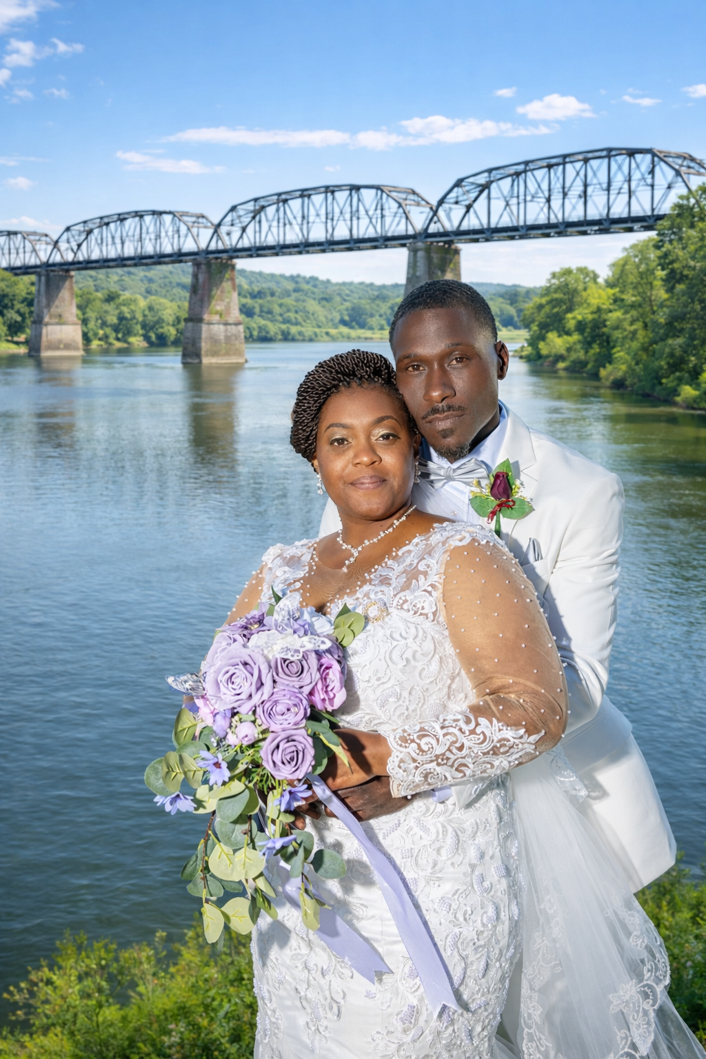 Bride and groom pose by a river with a steel bridge backdrop, she holds purple roses bouquet in white lace wedding dress.