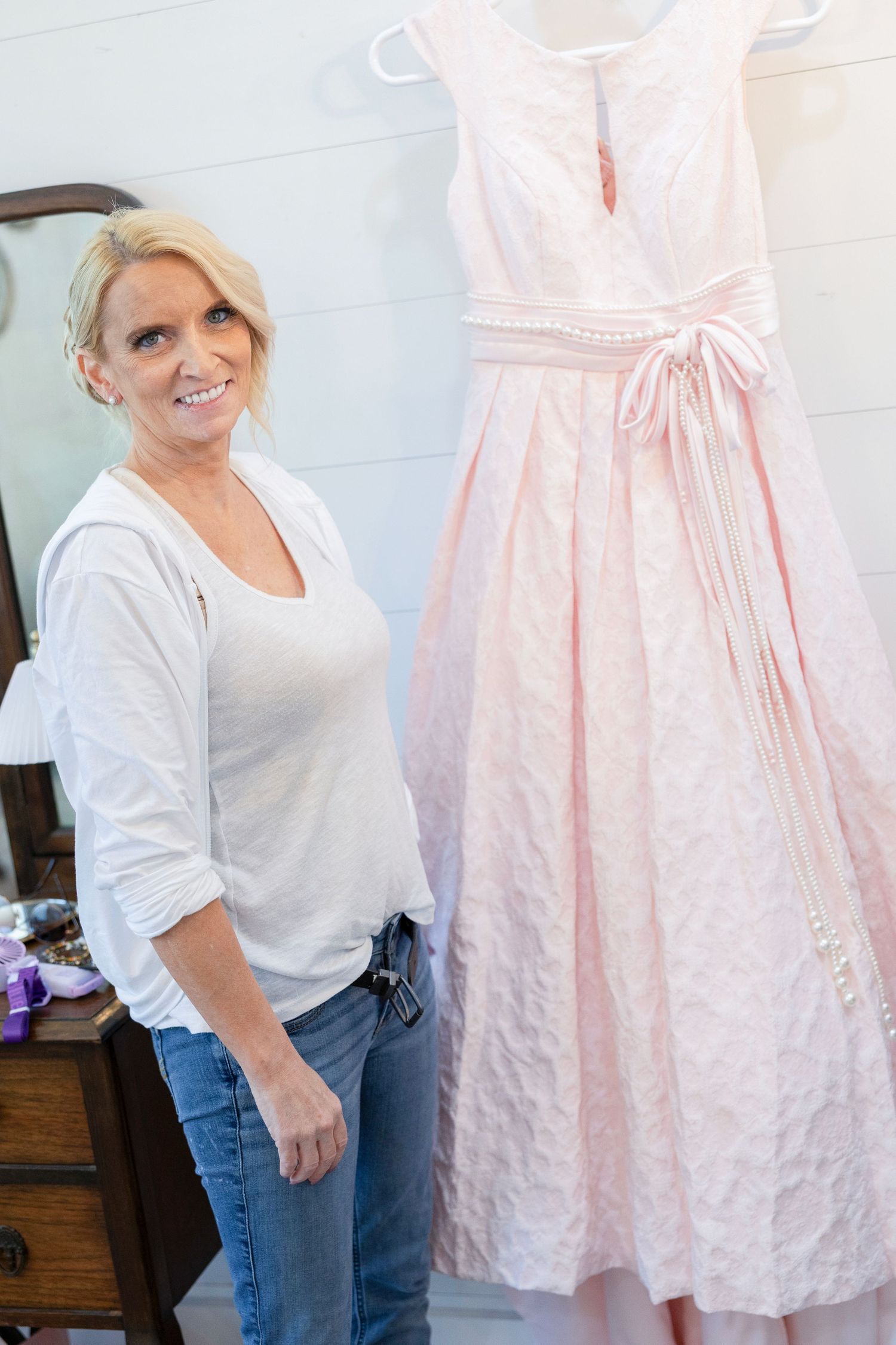 Smiling blonde woman in white top and jeans standing next to a pink vintage dress on a hanger.