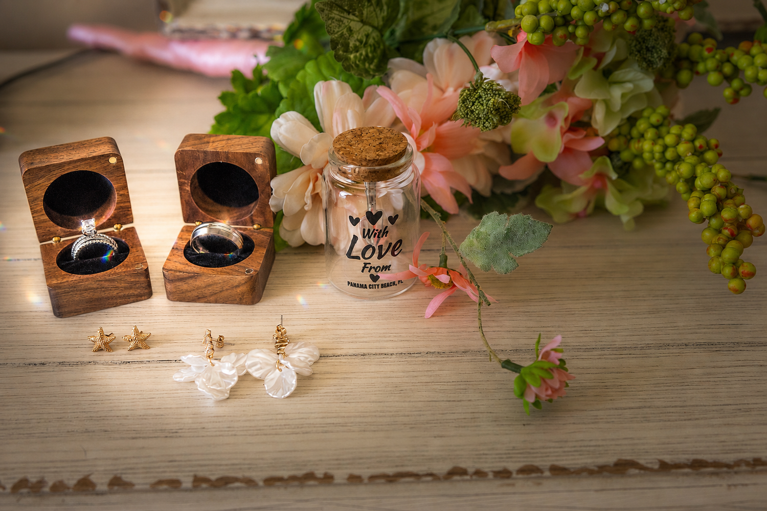 Wedding rings in wooden boxes displayed with love jar, floral bouquet, and delicate earrings on white surface.