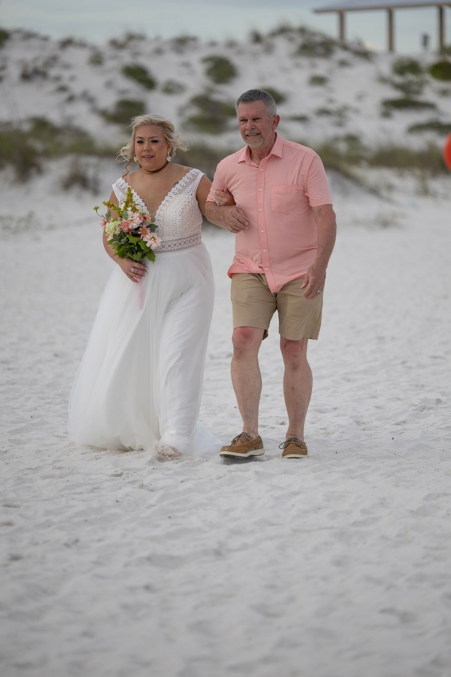 Bride in white gown holding bouquet walks with man in pink shirt on white sand beach wedding.