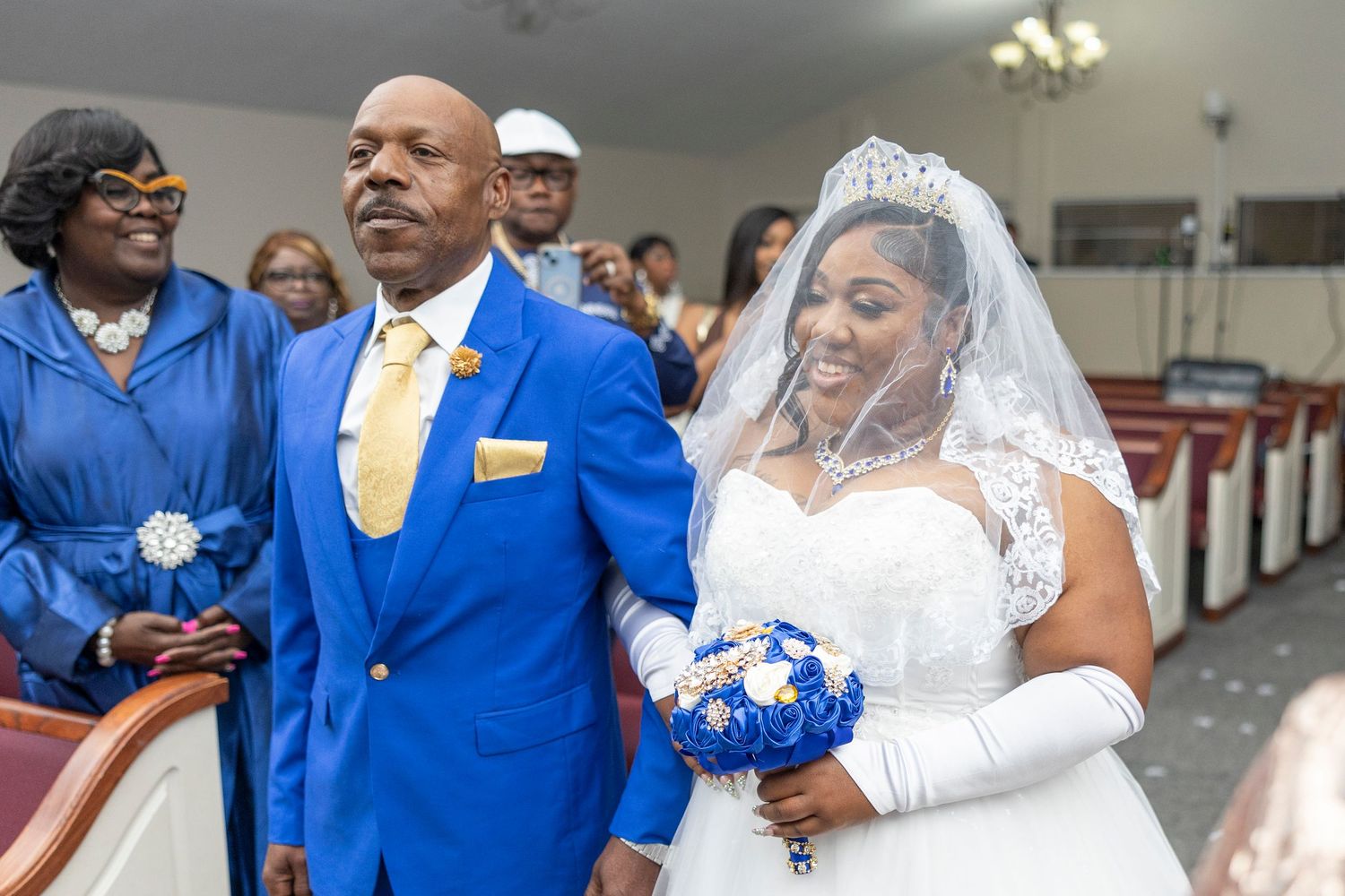 Bride in white gown and veil walks down church aisle with man in blue suit holding bouquet at wedding ceremony.