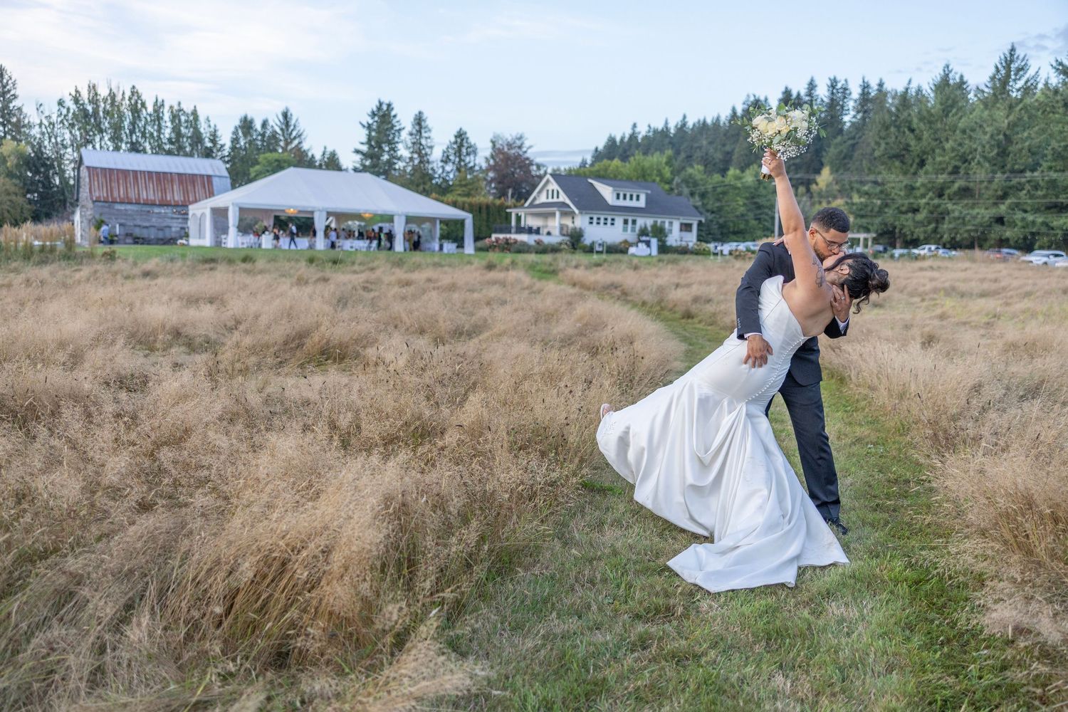 A romantic dip pose captured in a rustic field with tall golden grass and buildings in the background.