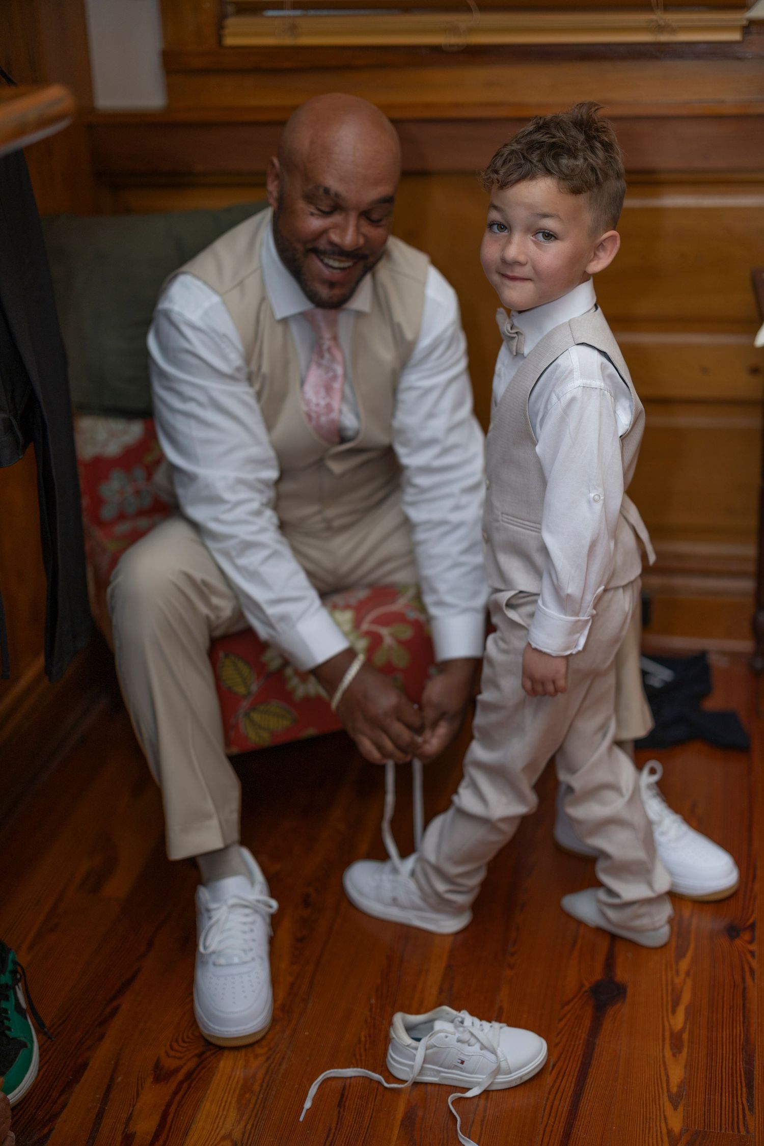 A heartwarming moment shows someone helping a small child tie their white dress shoes on a wooden floor.