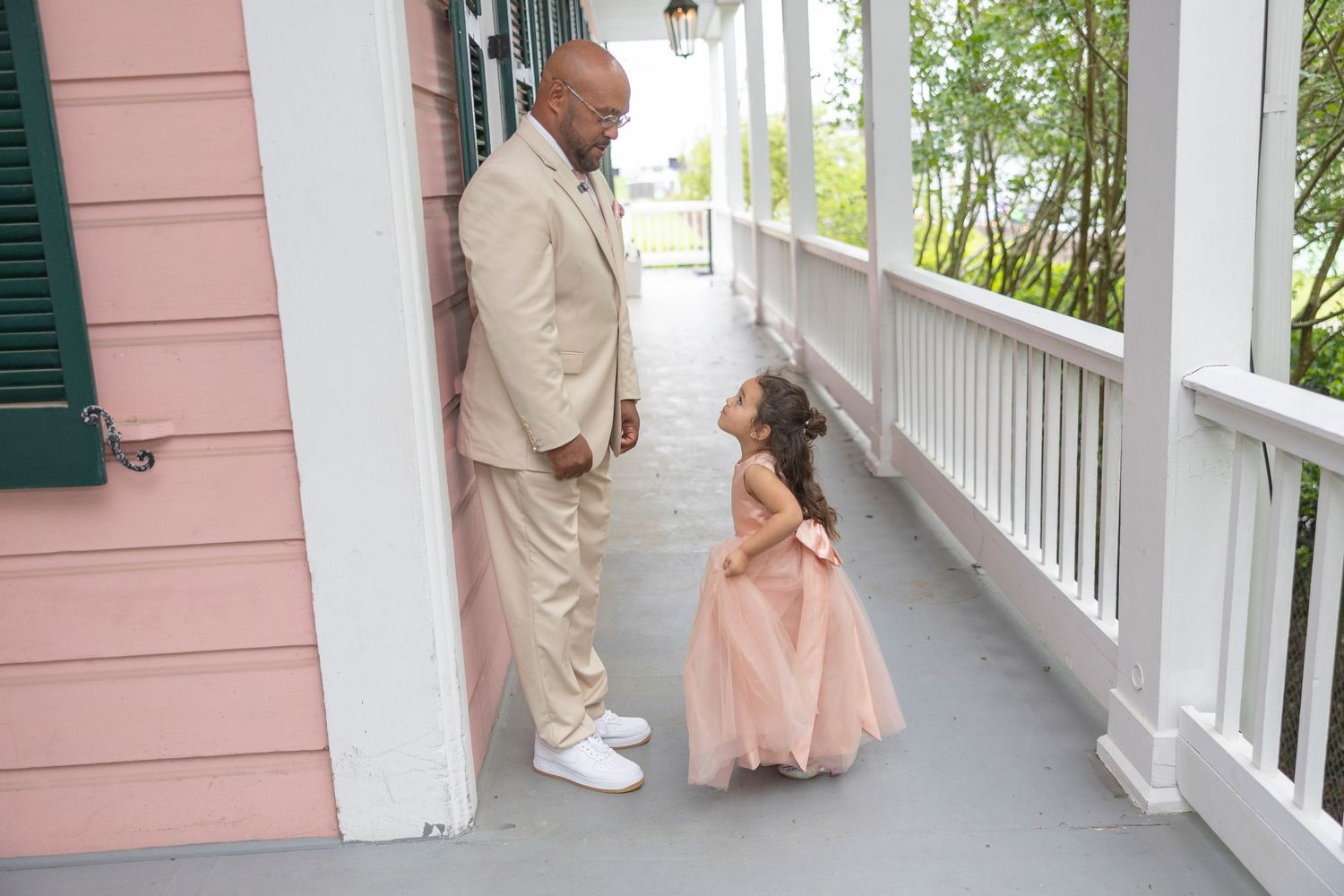 A wedding party poses on a covered porch of a pink house with white columns and green shutters.