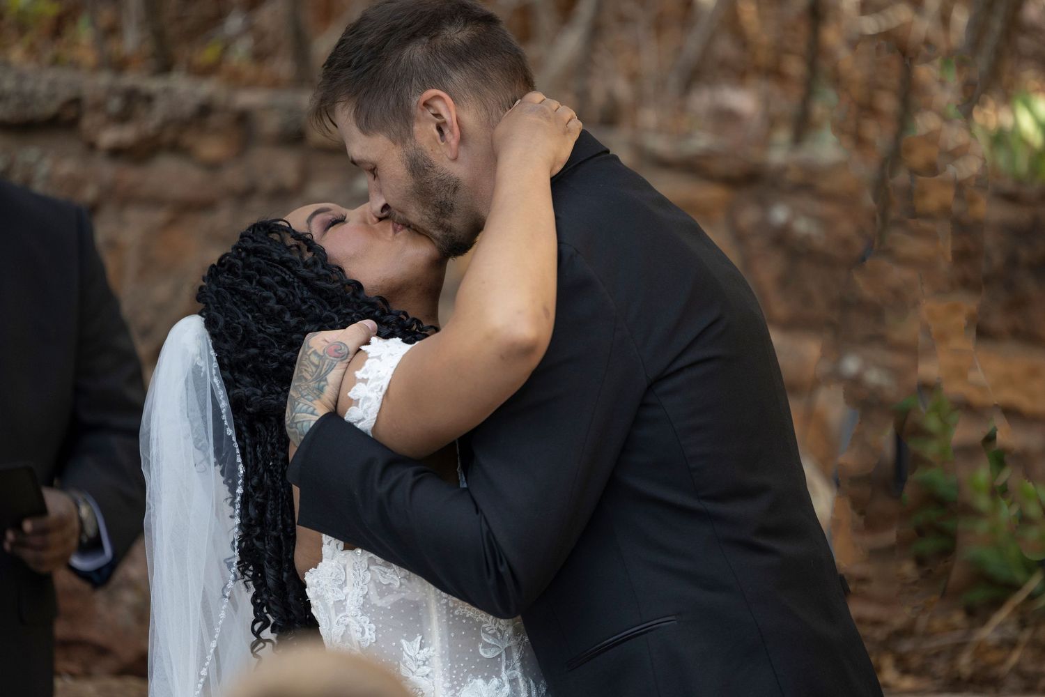 A romantic couple shares a tender embrace and kiss in front of a rustic brick wall background.