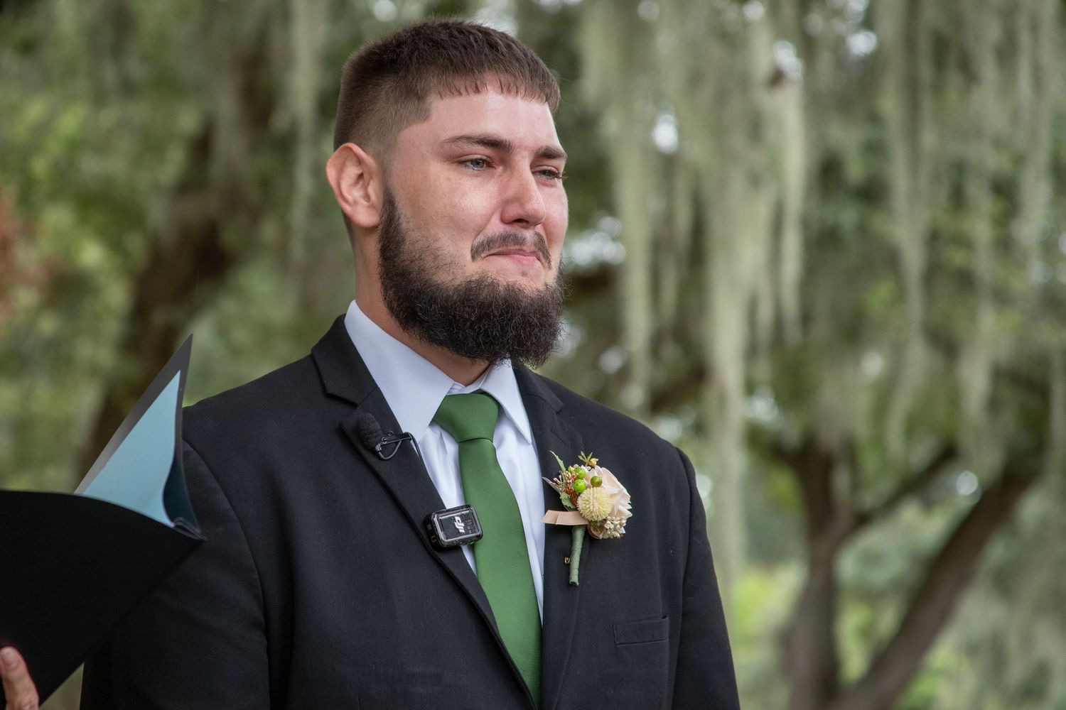 A groom in a black suit with green tie stands outdoors among trees during a wedding ceremony.