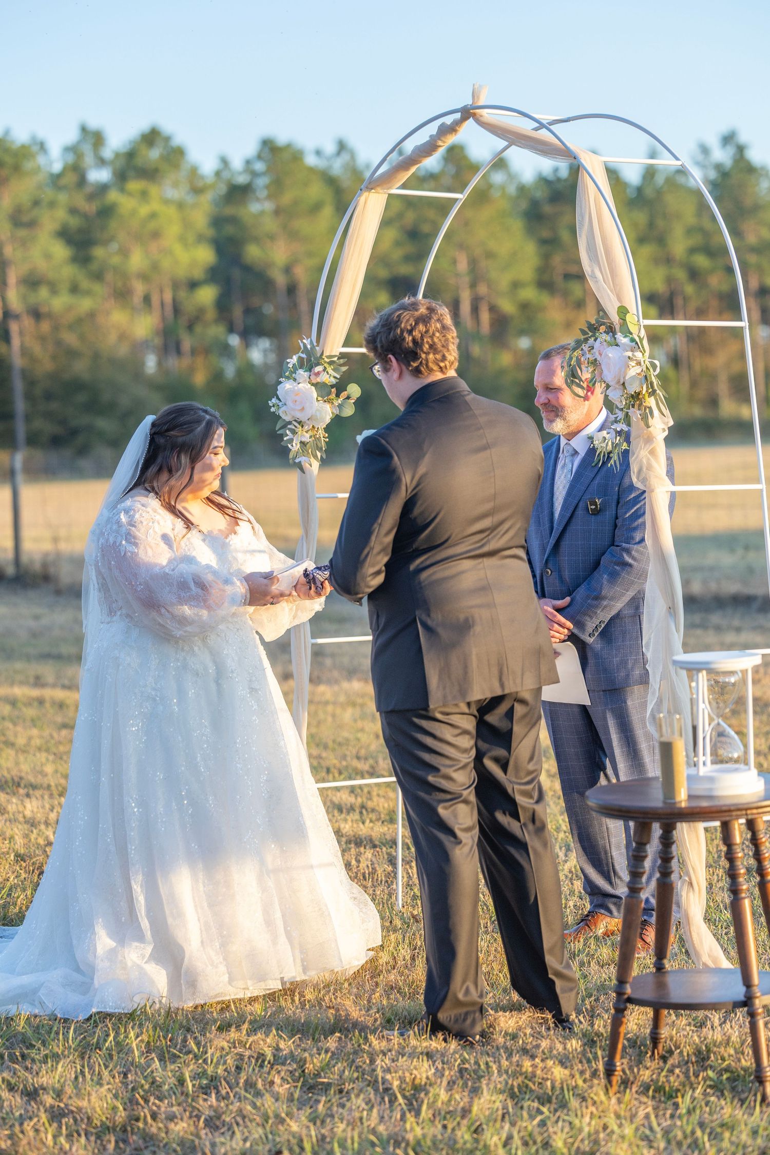 Outdoor wedding ceremony at sunset with couple and officiant standing by decorative white arch in grassy field.
