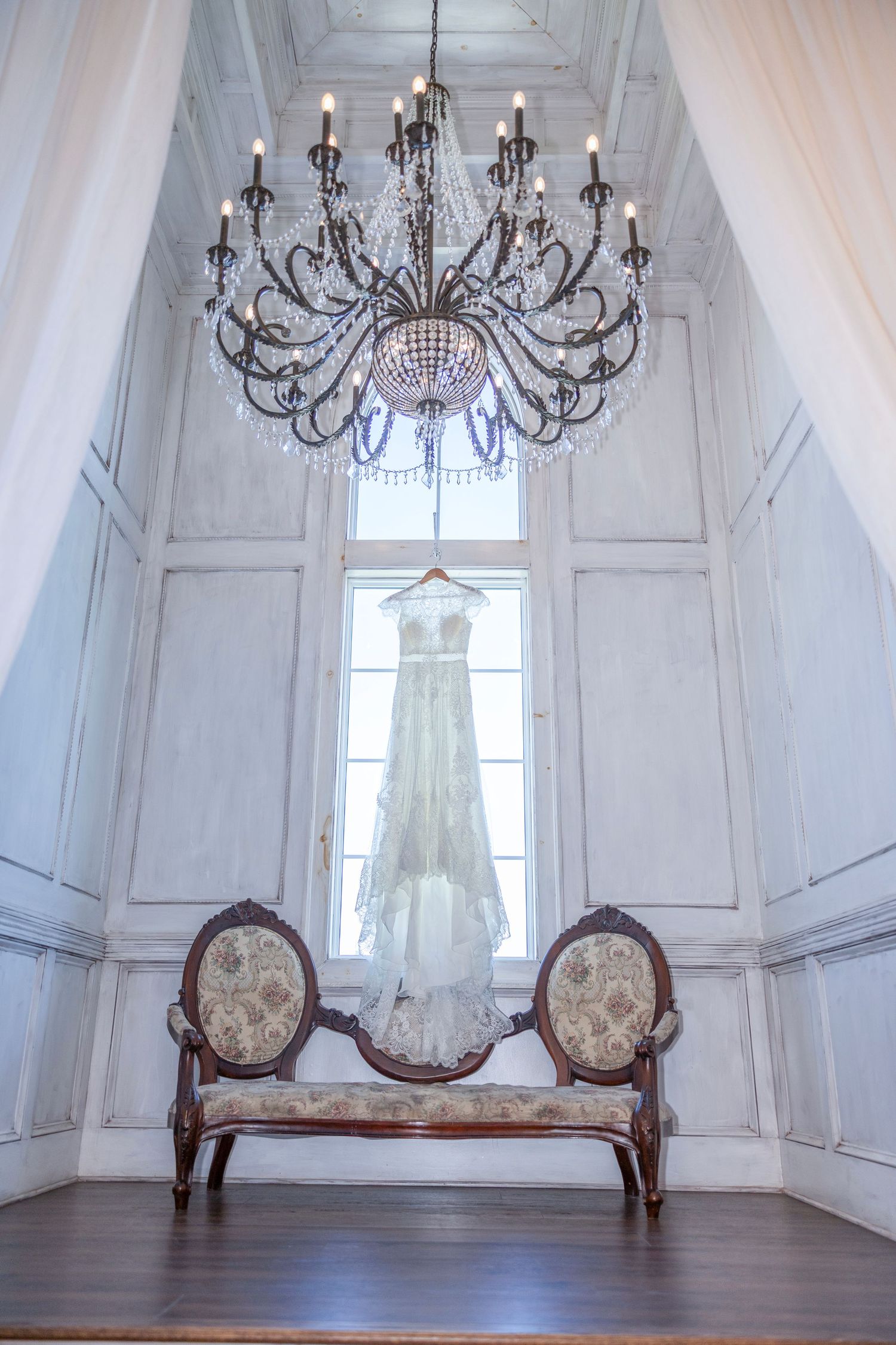 Elegant wedding dress hangs in tall white room with ornate chandelier and vintage settee beneath window.