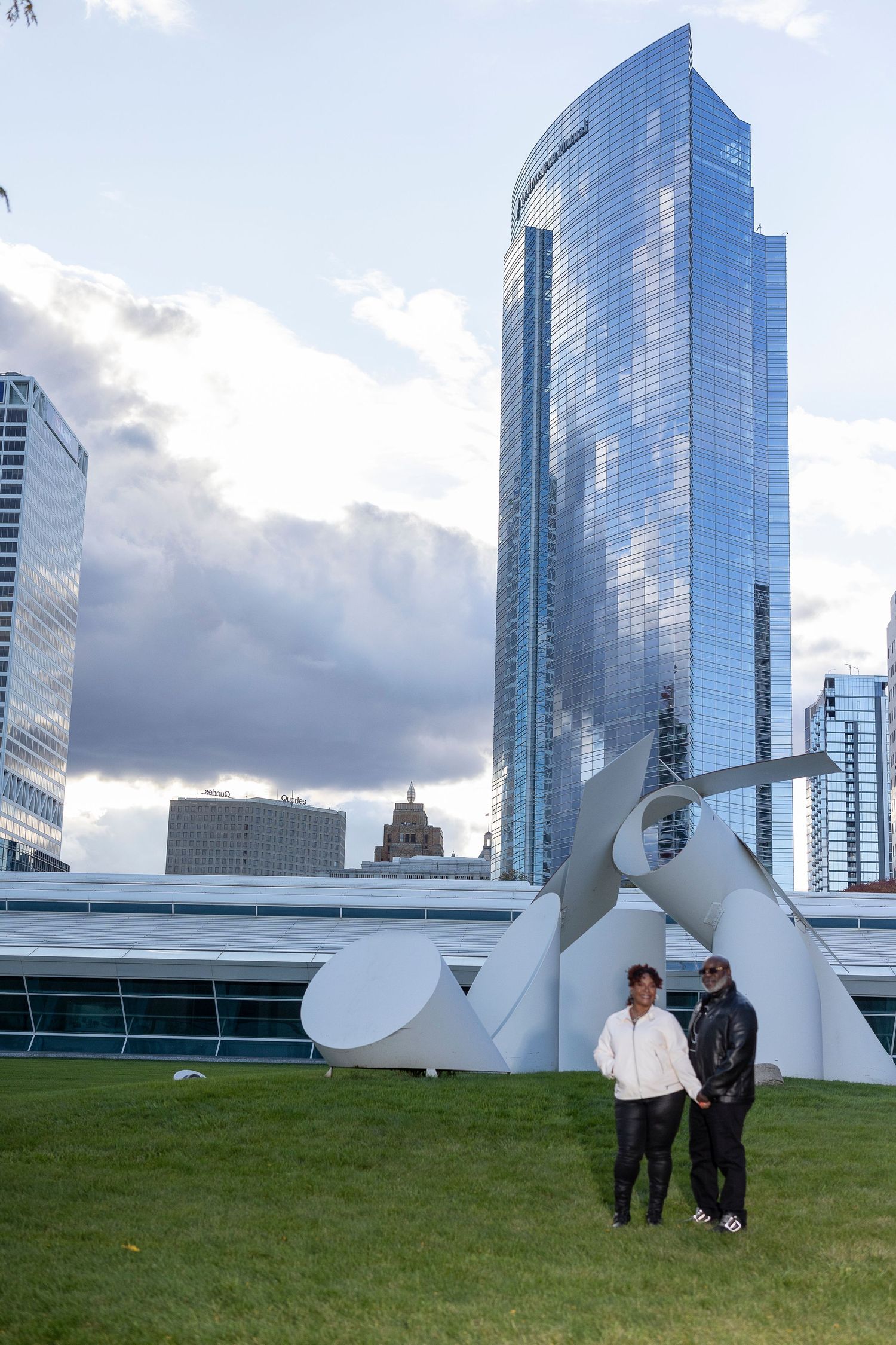 Modern art sculpture with curved white forms sits on grassy hill against backdrop of glass skyscrapers in urban setting.