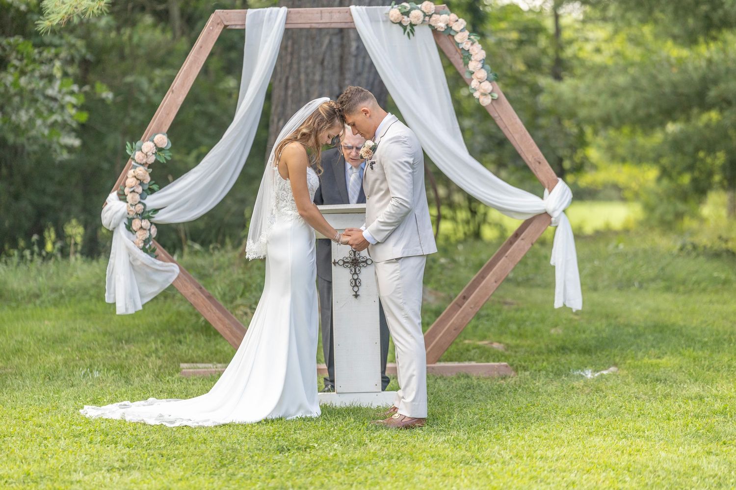 A couple stands under a wooden arch decorated with white fabric and floral arrangements during an outdoor wedding ceremony.