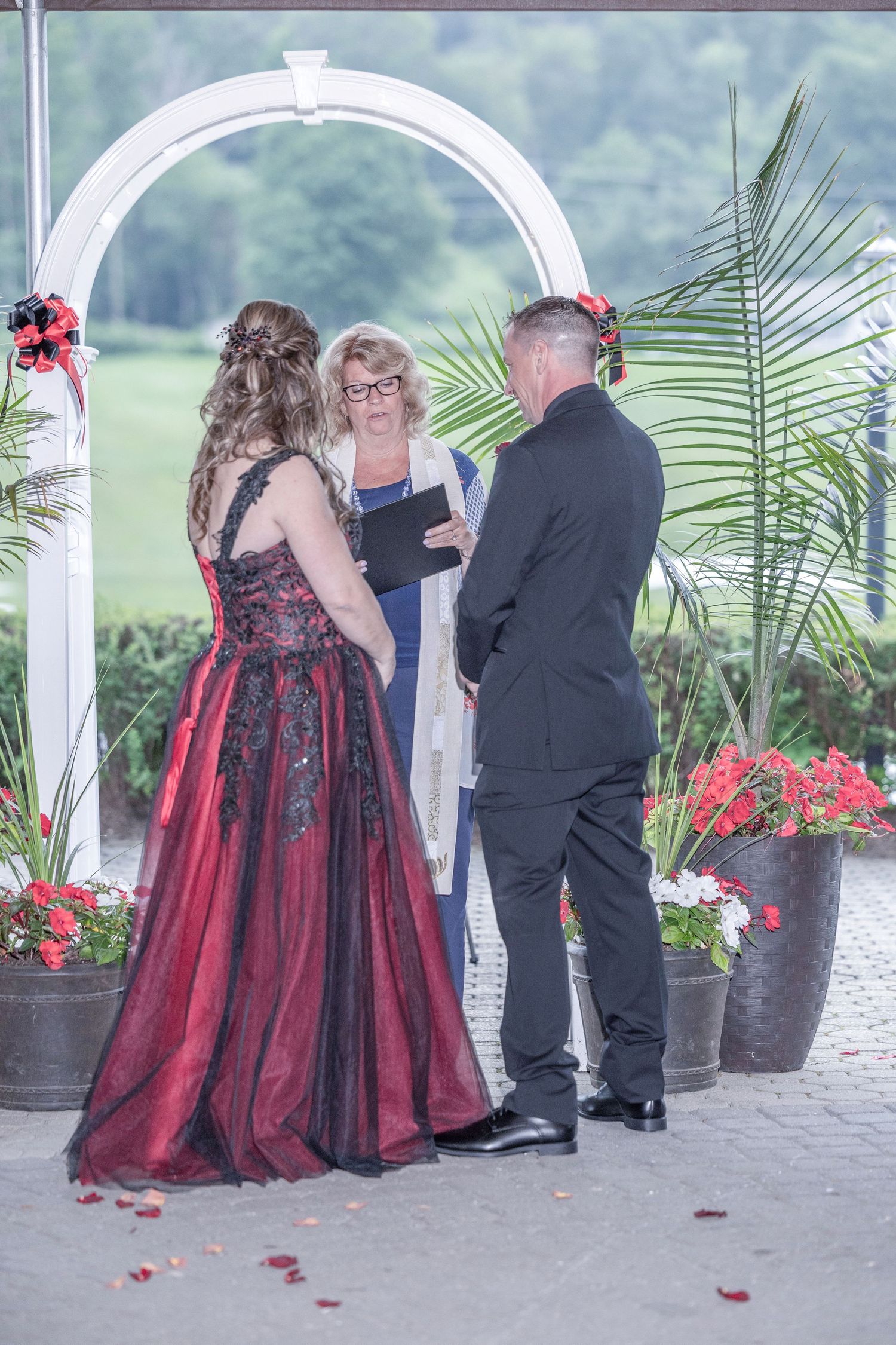 Wedding party gathers under white gazebo decorated with red flowers and palm plants during outdoor ceremony.
