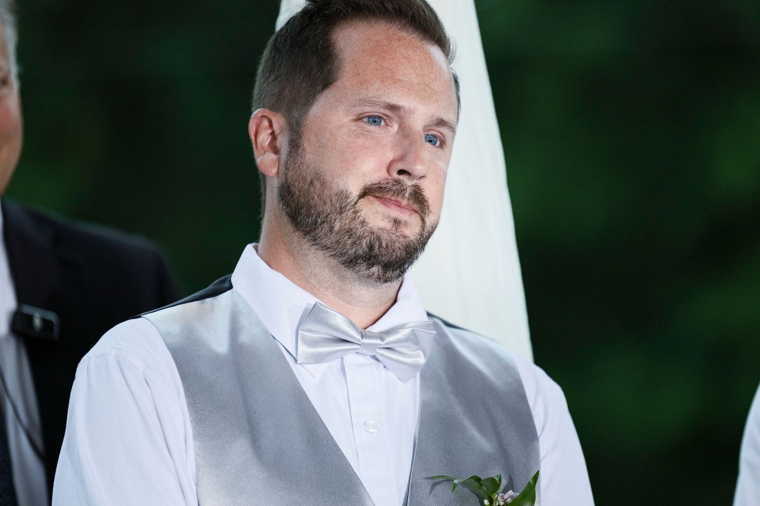 A groom in a white dress shirt and gray vest stands outdoors against a blurred green natural background.
