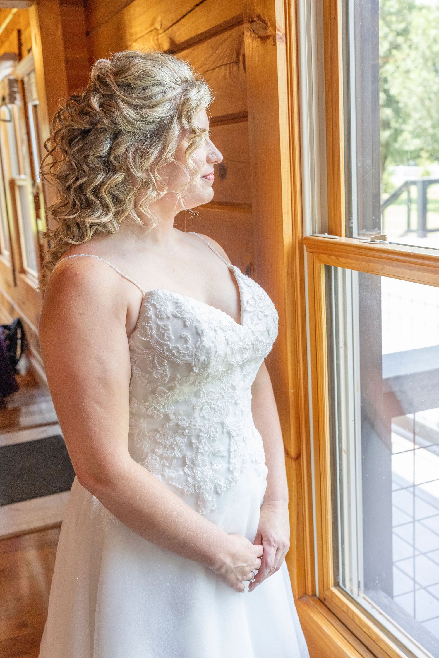 A bride in a white lace wedding dress with curled hair stands by a window in a rustic wooden room.