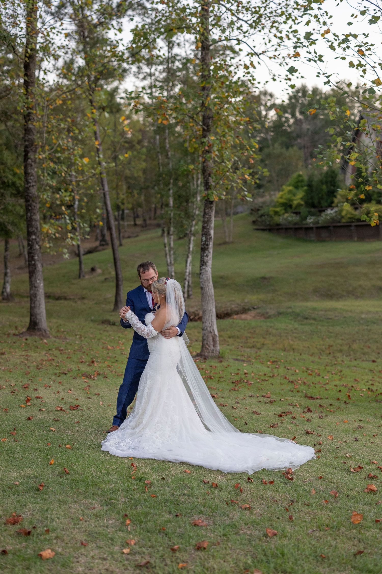 A couple shares romantic moments in a wooded park setting with scattered autumn leaves on the ground during their wedding day.