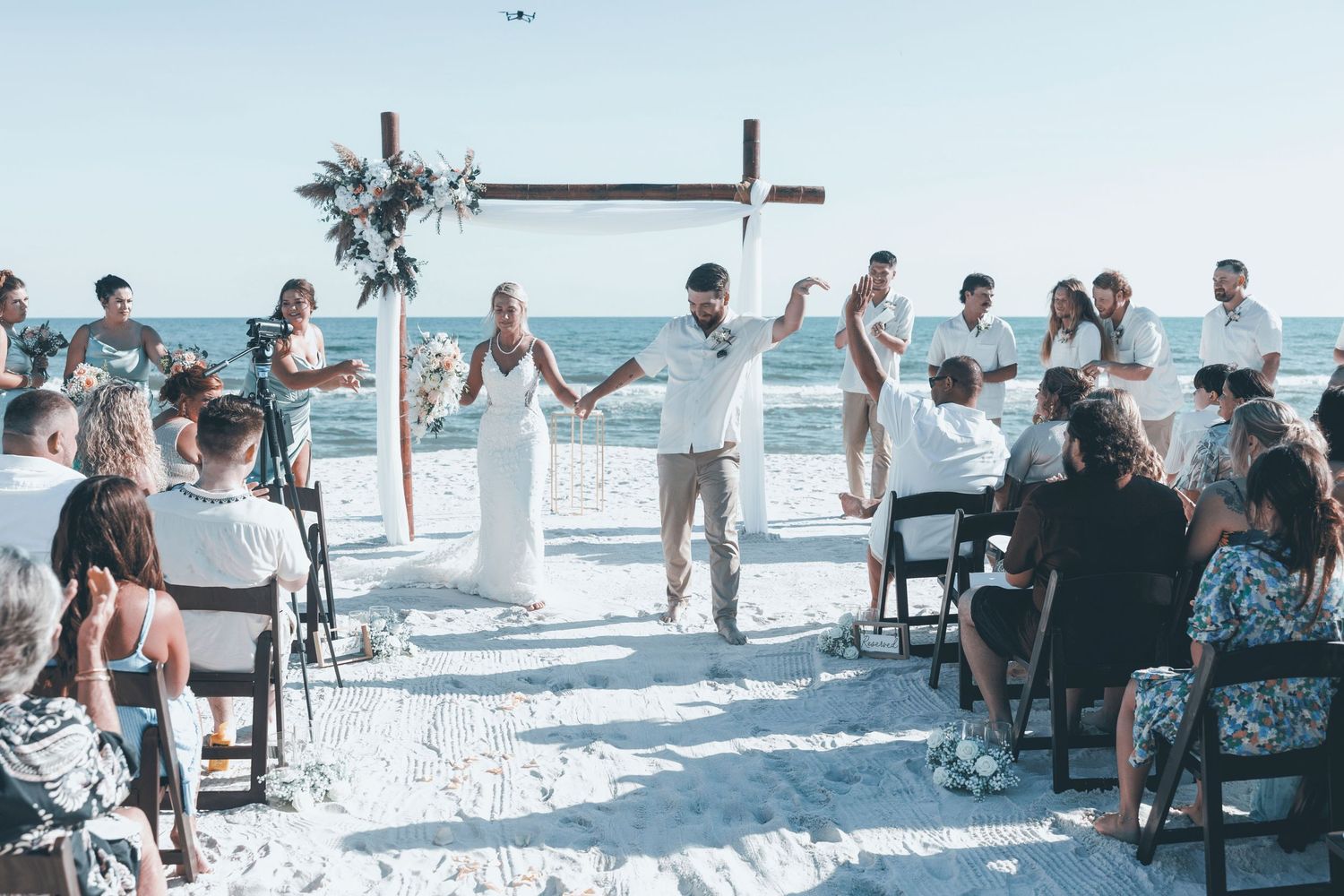 A beachside wedding ceremony with guests seated on chairs in the sand as a couple celebrates at a wooden cross altar.