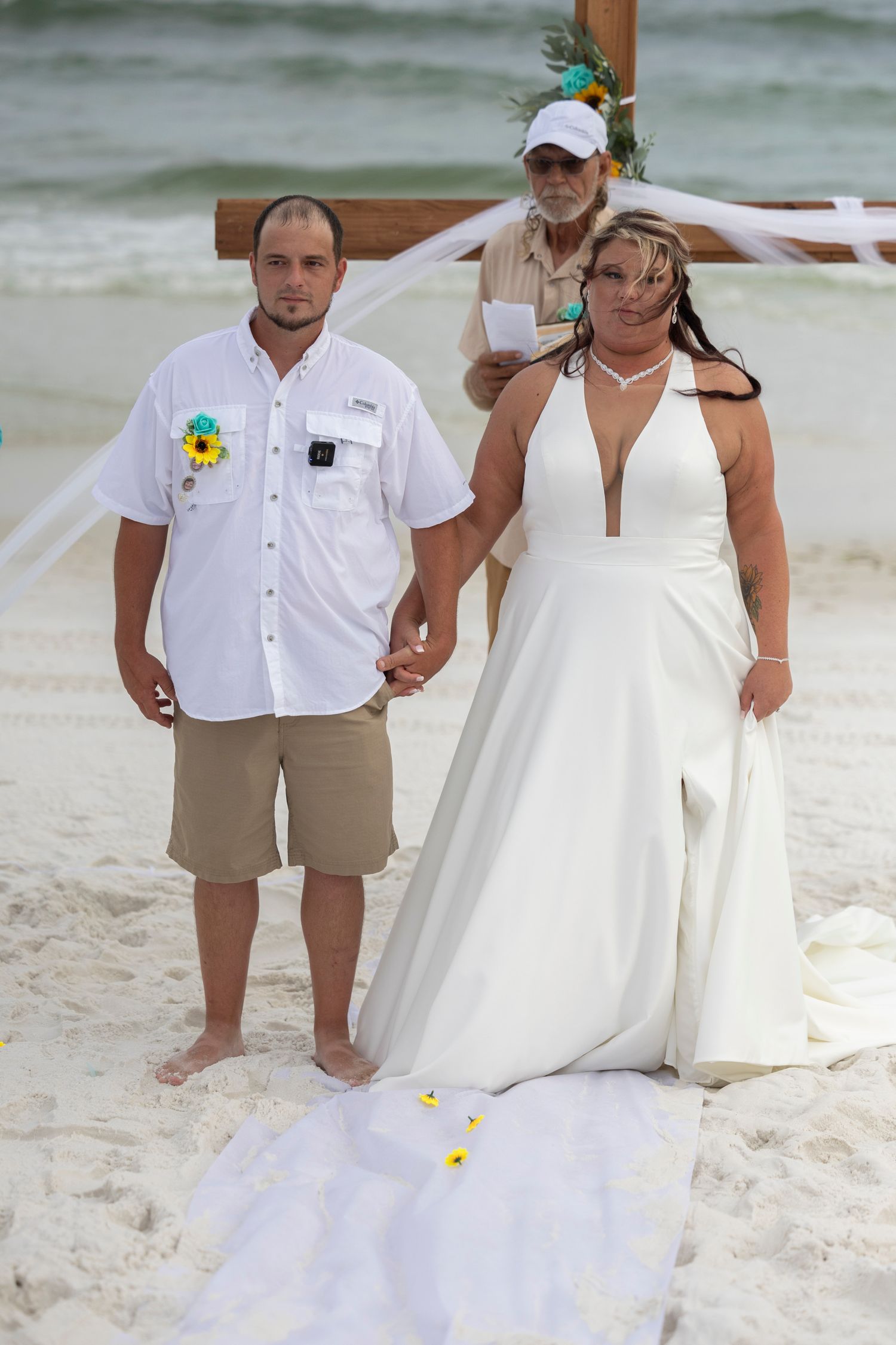 A couple in wedding attire poses on a white sandy beach with ocean waves in the background.