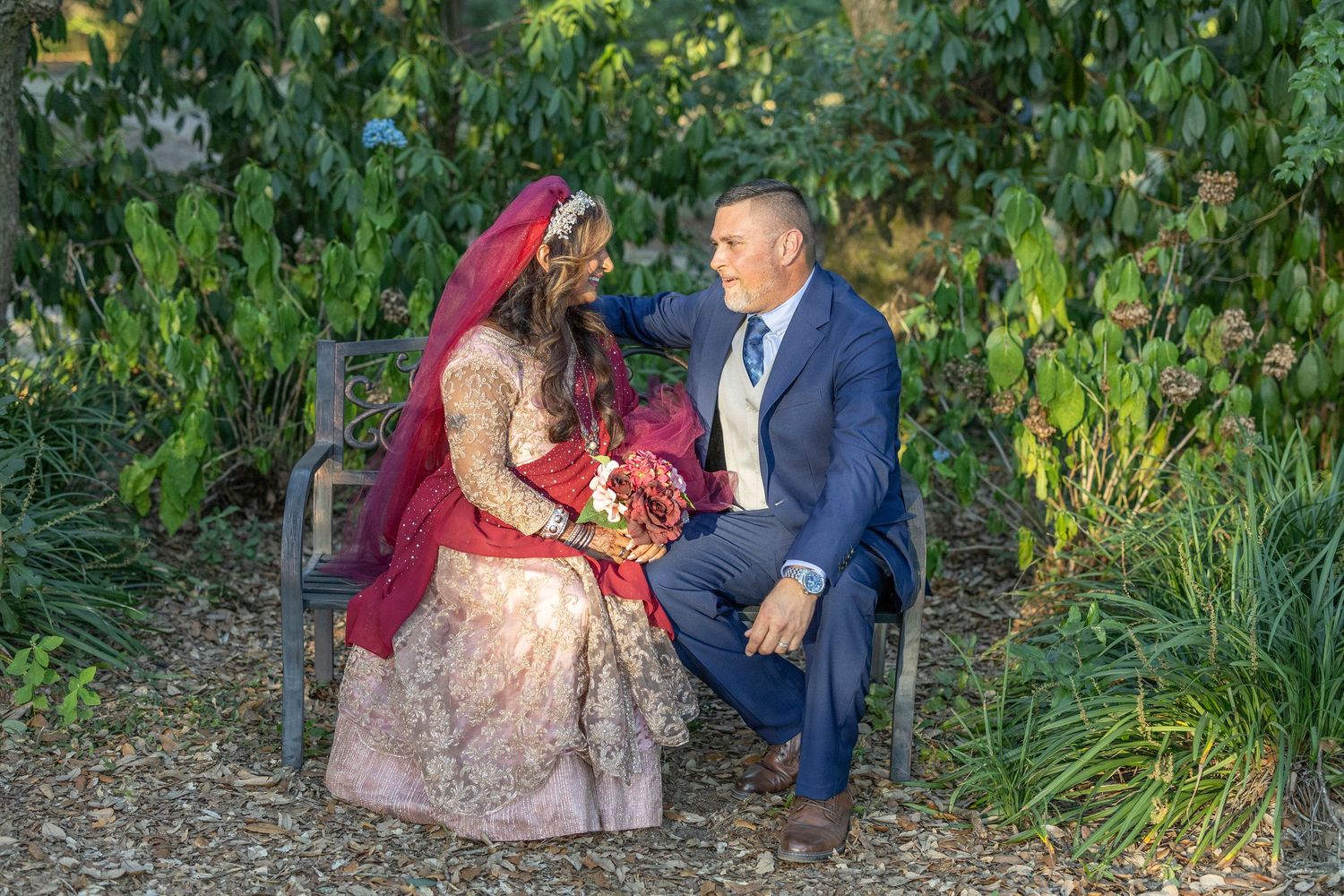 A couple in formal attire poses together in a lush garden setting during a romantic outdoor photoshoot.