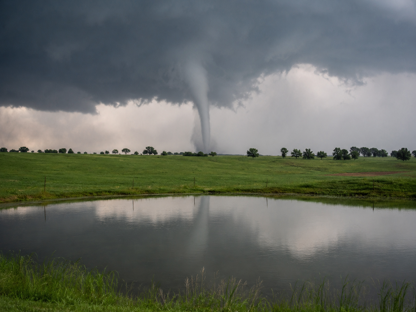 A large tornado reflected in a calm pond surrounded by lush green fields under stormy skies.