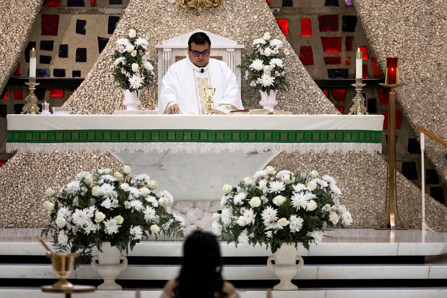 Catholic priest celebrates Mass at ornate white altar adorned with white floral arrangements in a decorated church.