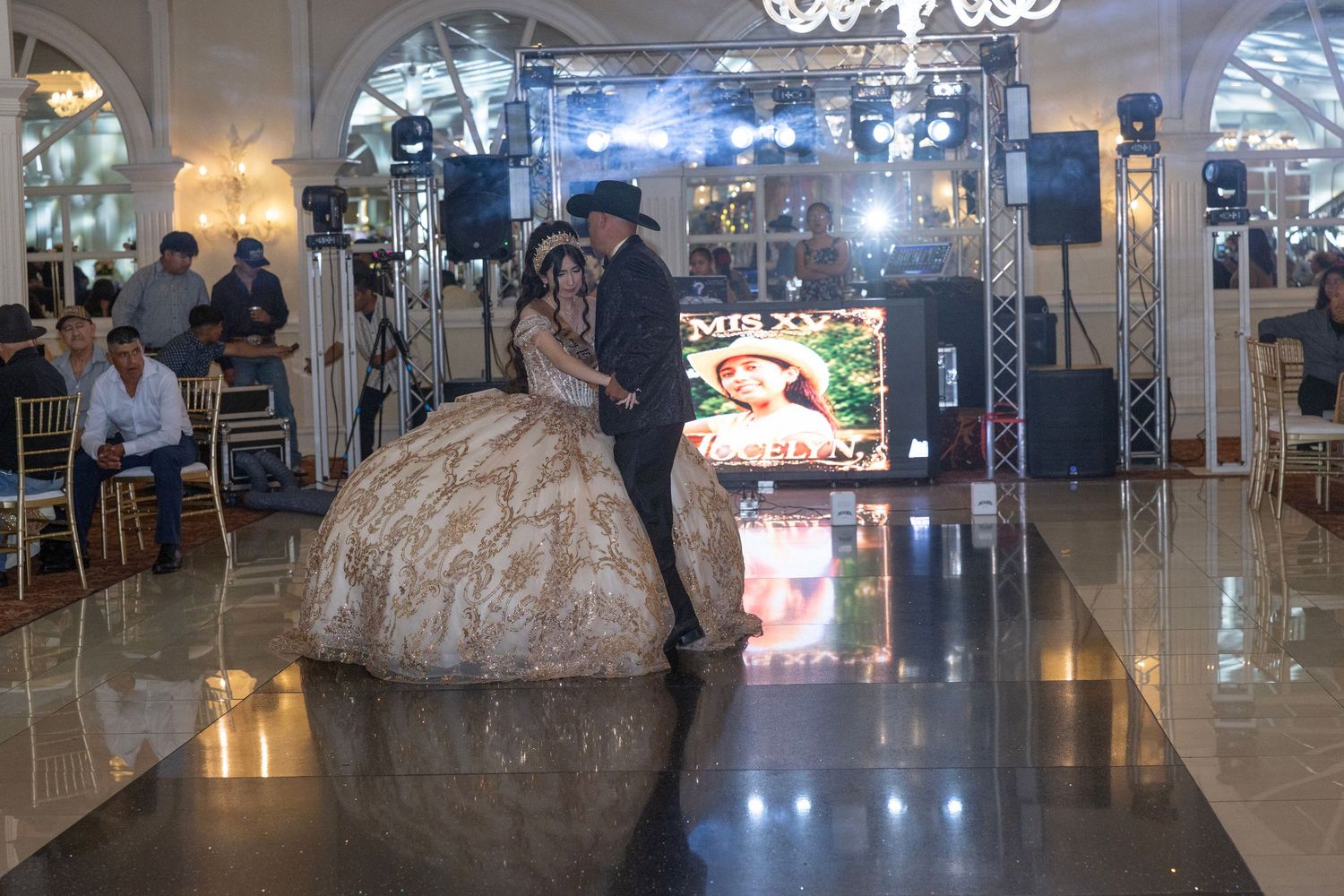A young woman in a gold quinceañera gown shares a dance with a partner at an elegant ballroom celebration.