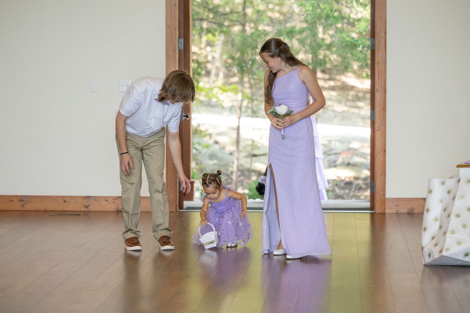 A boy and bridesmaid in lavender gown interact with a young flower girl in purple dress on a wooden dance floor.