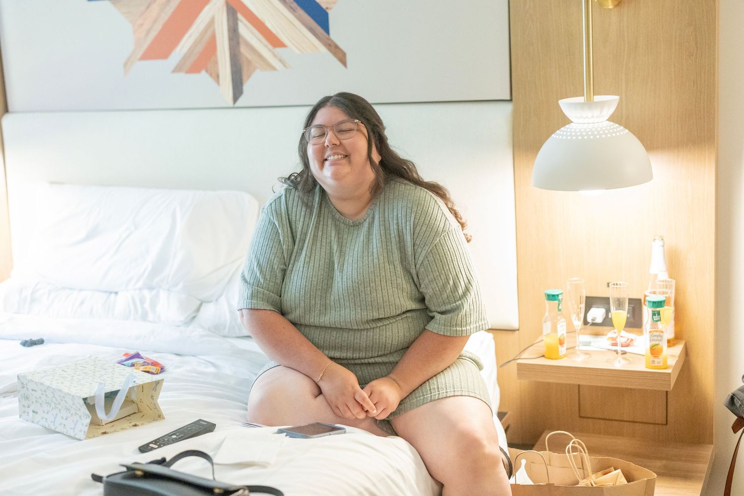 Plus-size woman smiling while sitting on a white hotel bed next to a nightstand with toiletries.