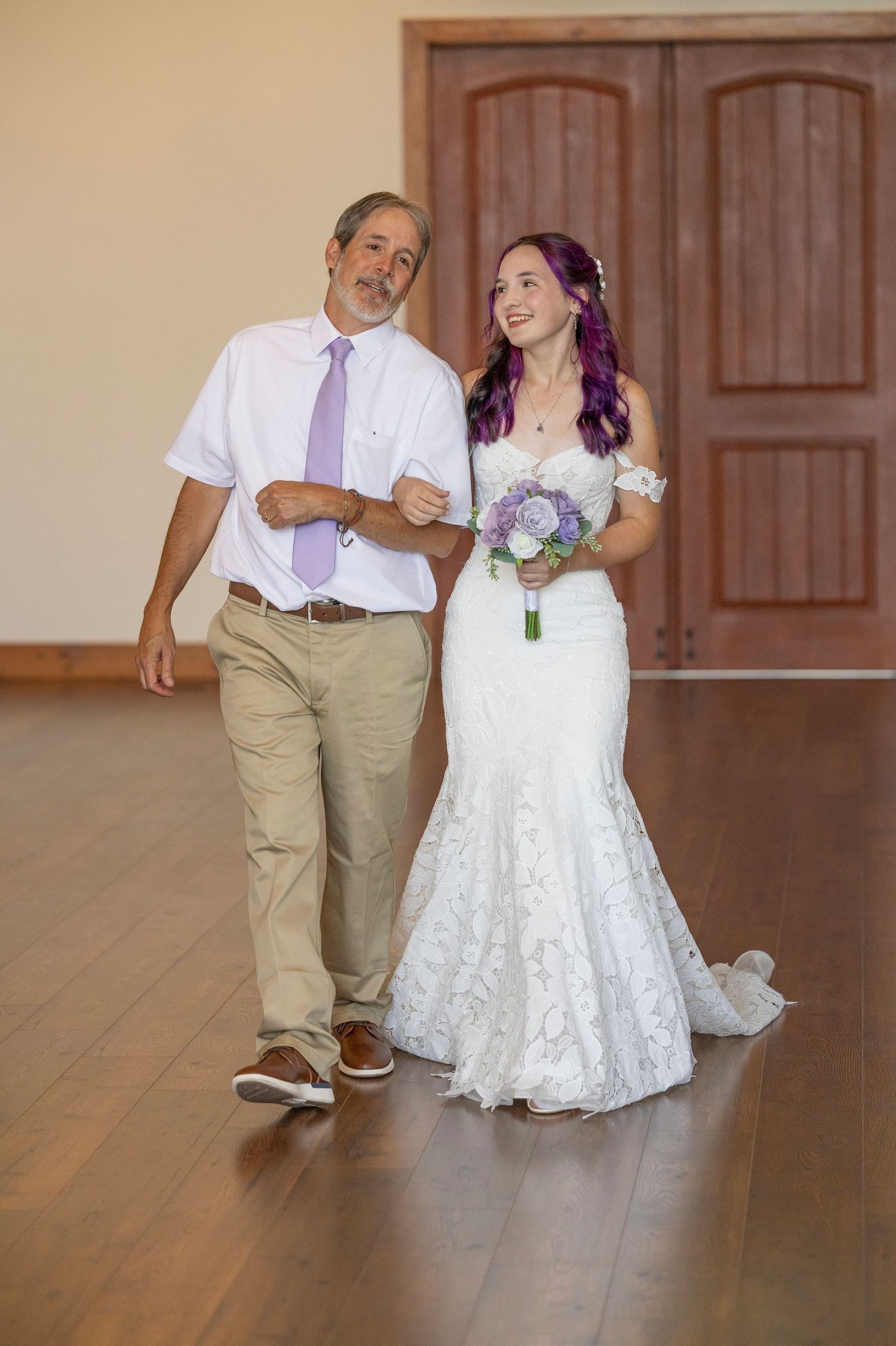 Bride in white lace gown with purple bouquet walks with older man in lavender tie on wedding day.