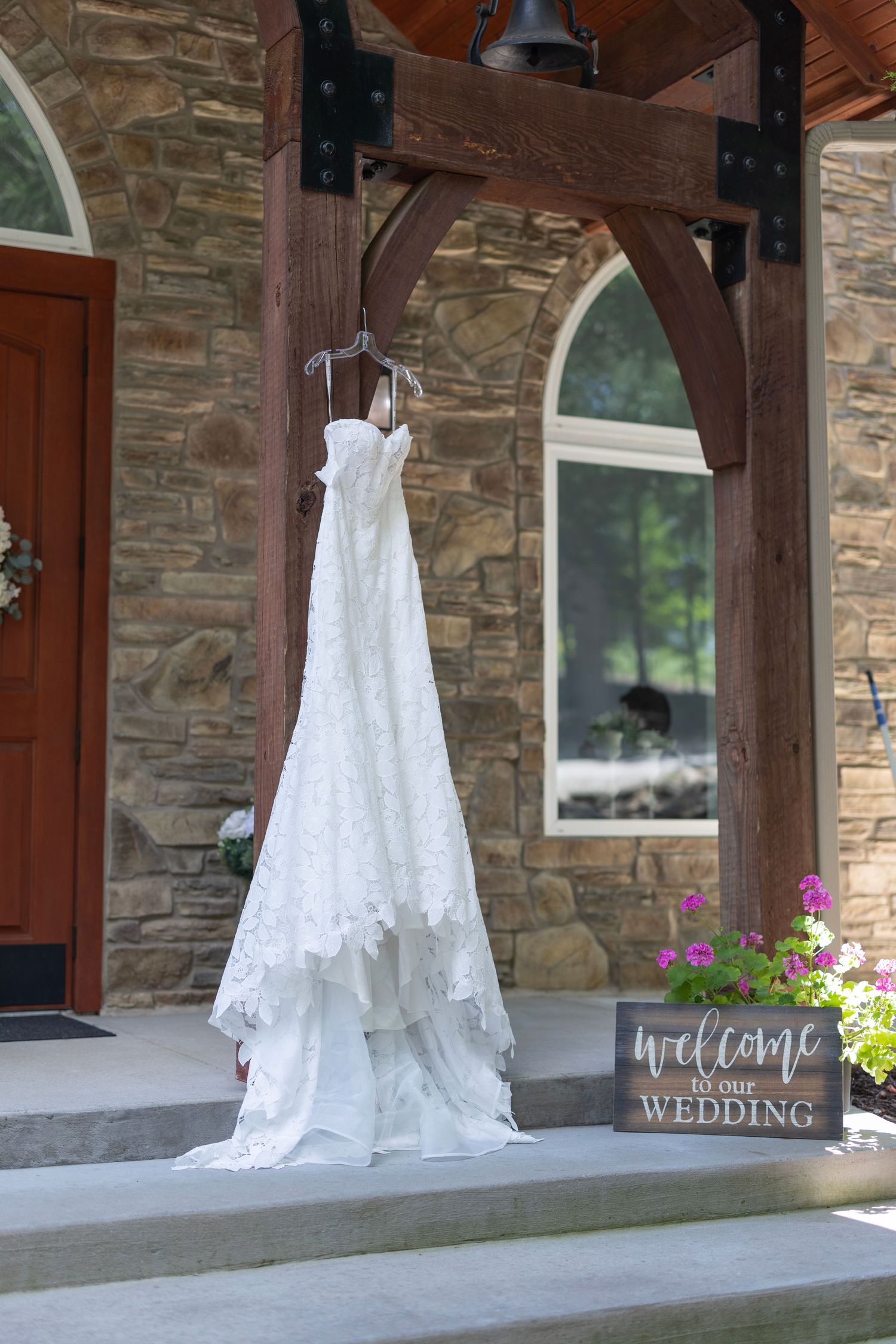 White lace wedding dress hanging on a wooden beam outside a stone chapel with a Welcome to Our Wedding sign.